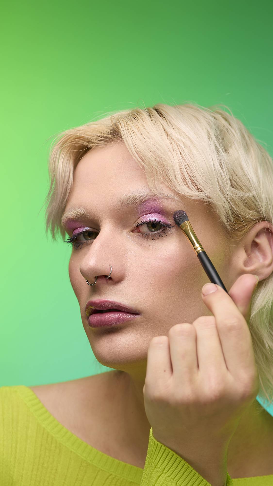The image shows a close-up of the model's face on a pale blue-to-green gradient background. They are holding a thin make-up brush to apply a subtle, rose-pink, shimmering eyeshadow of the Big Bang Glitter Crumble. 