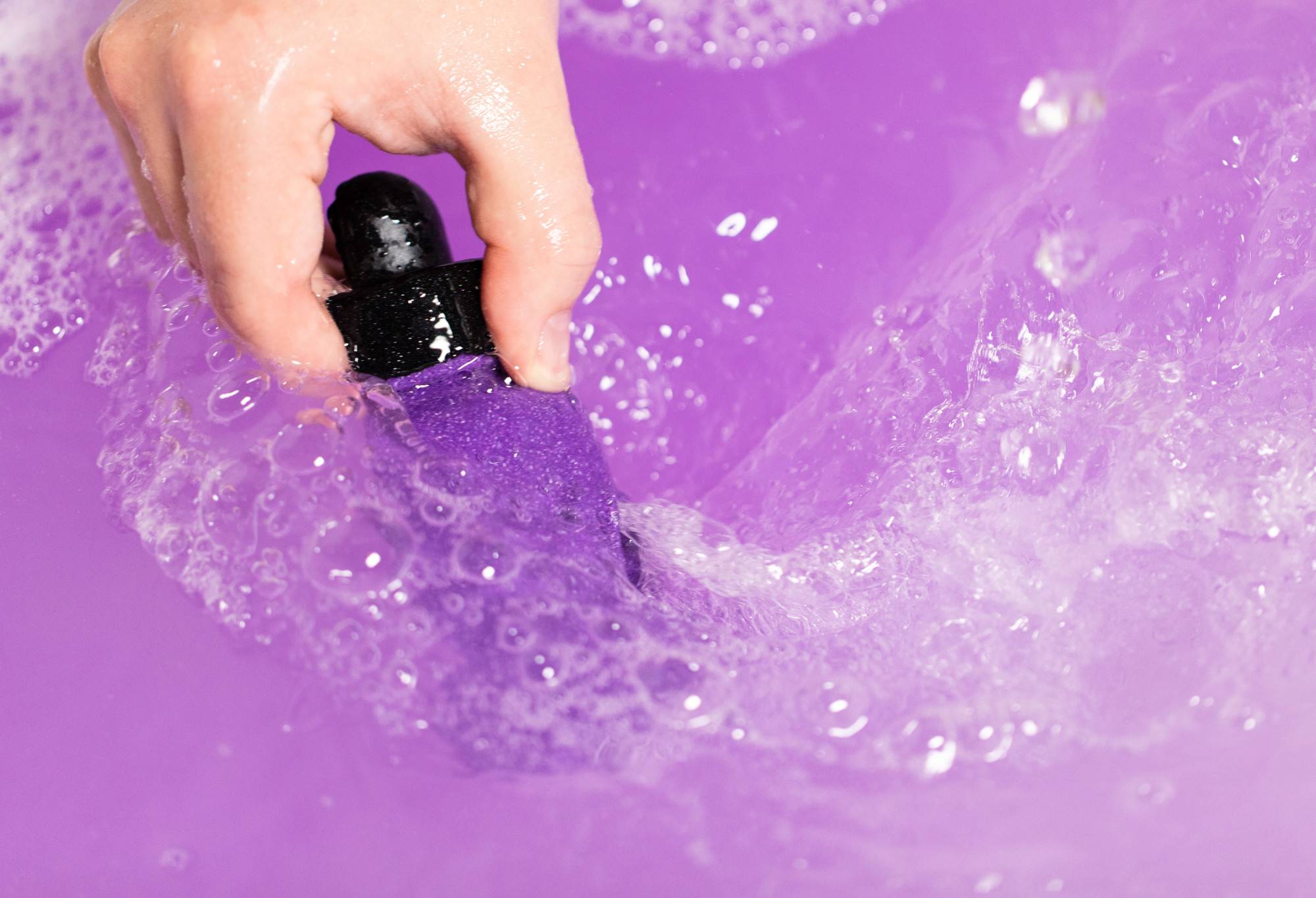 A purple, bottle shaped bubble bar is held by its black wax lid to swirl into bubbly water of a vibrant purple colour.