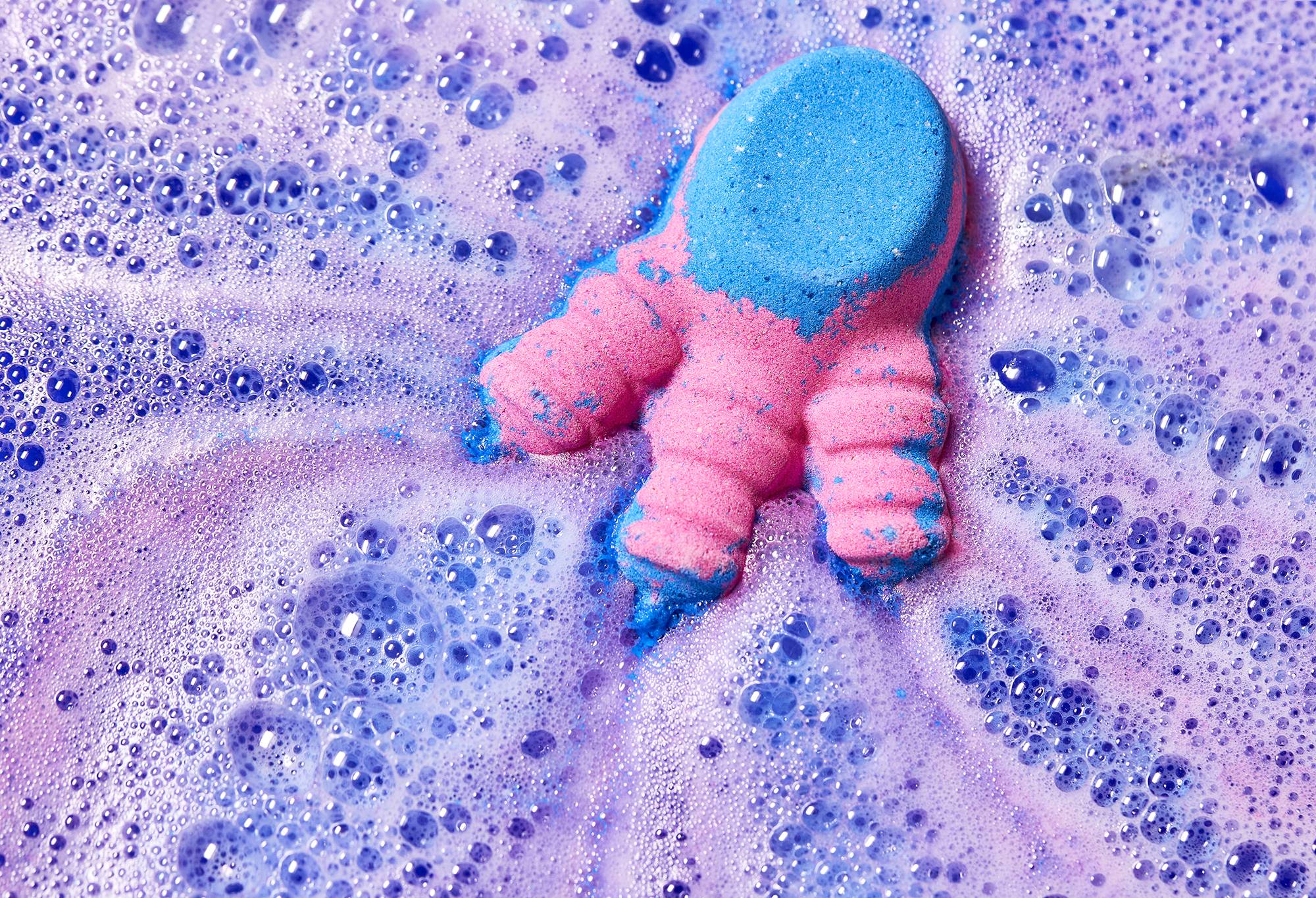 The bath bomb fizzes in the water, releasing blue and pink foam trails across the surface.