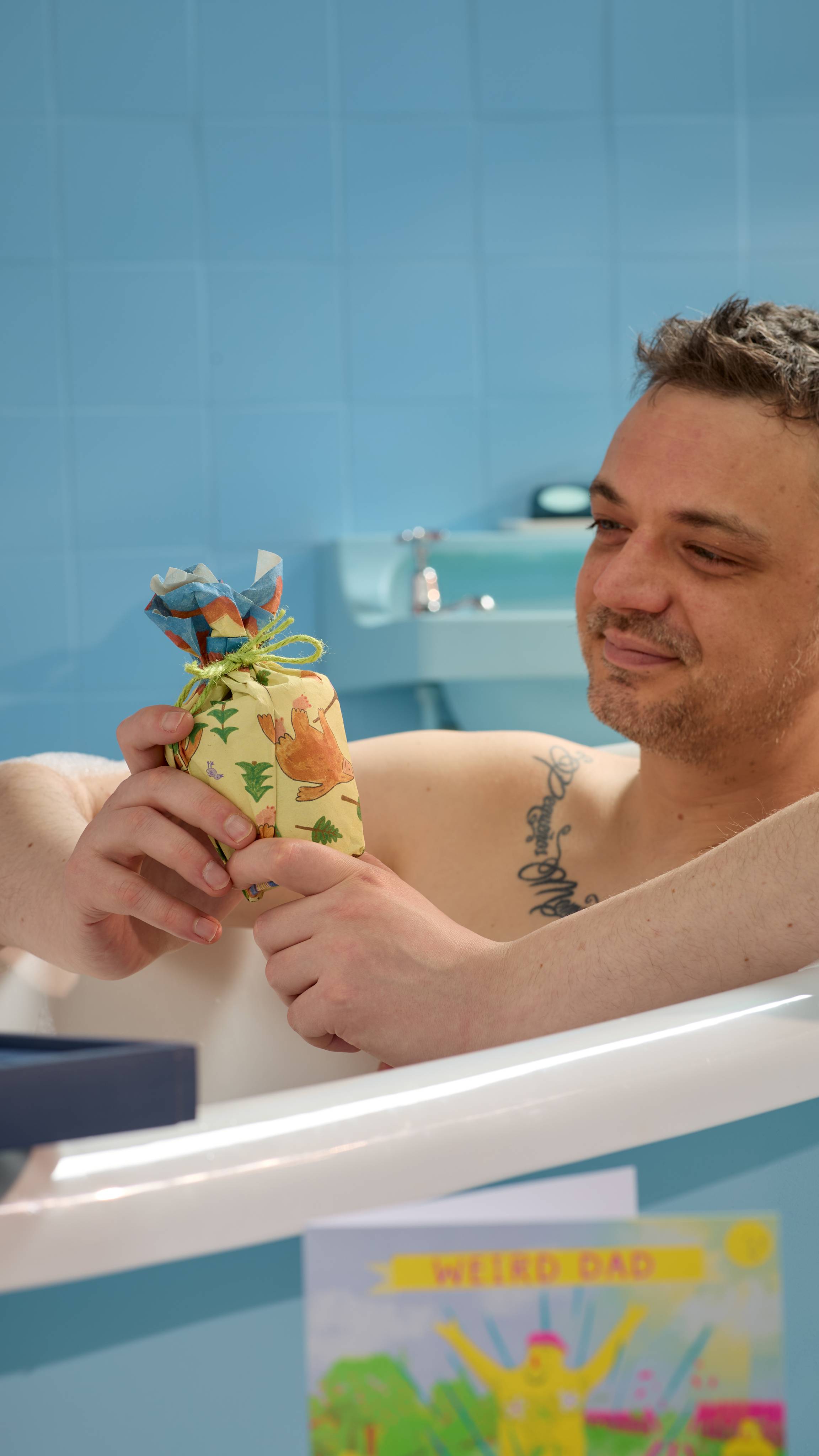 The Model is sitting in a roll-top bathtub in a blue, tiled room holding a gift wrapped in the Big Foot Goes Camping lokta wrap and tied with green string. 