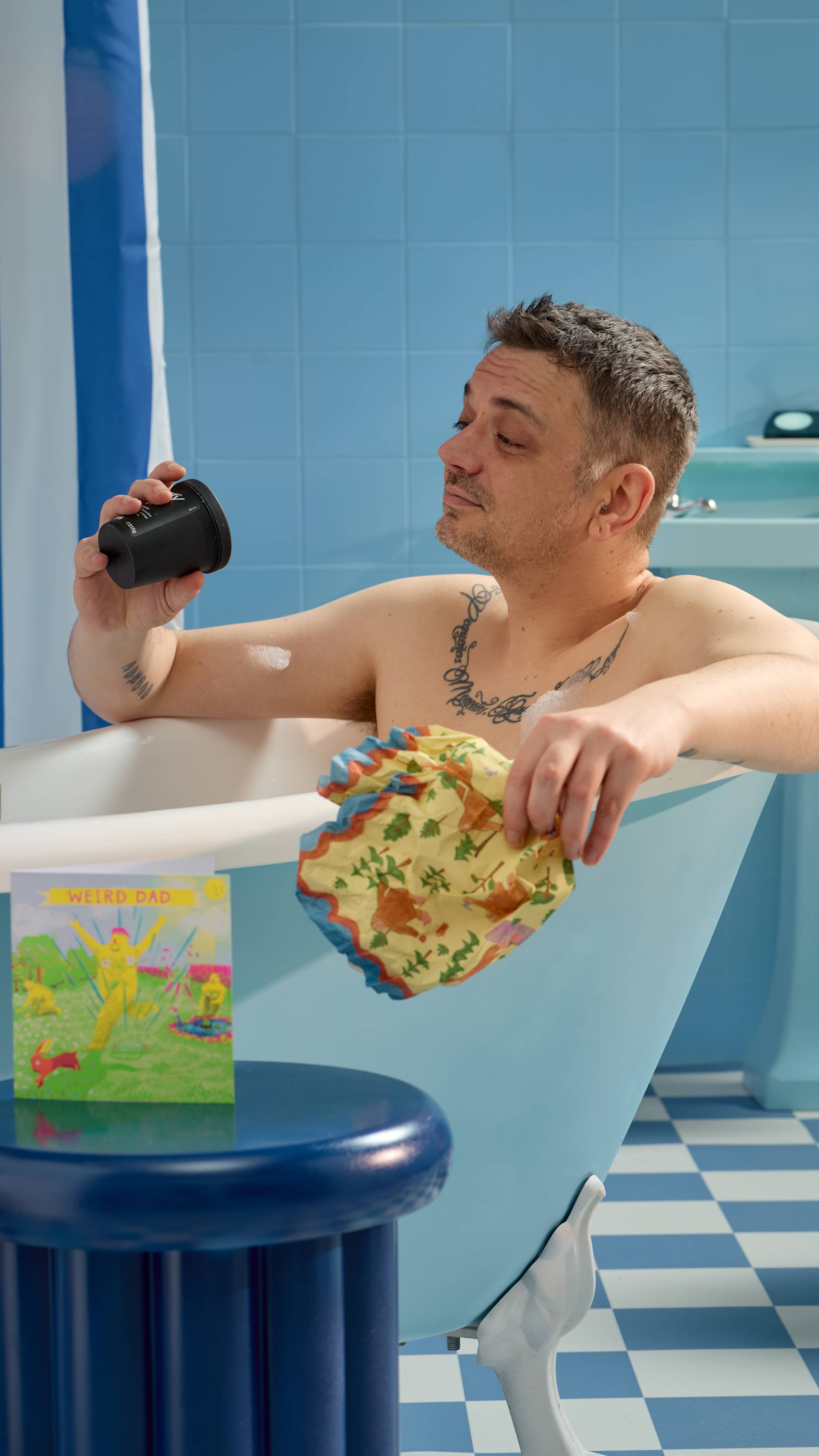 The Model is sitting in a roll-top bathtub in a blue, tiled room and has unwrapped a potted Lush product from the Big Foot Goes Camping lokta wrap. 