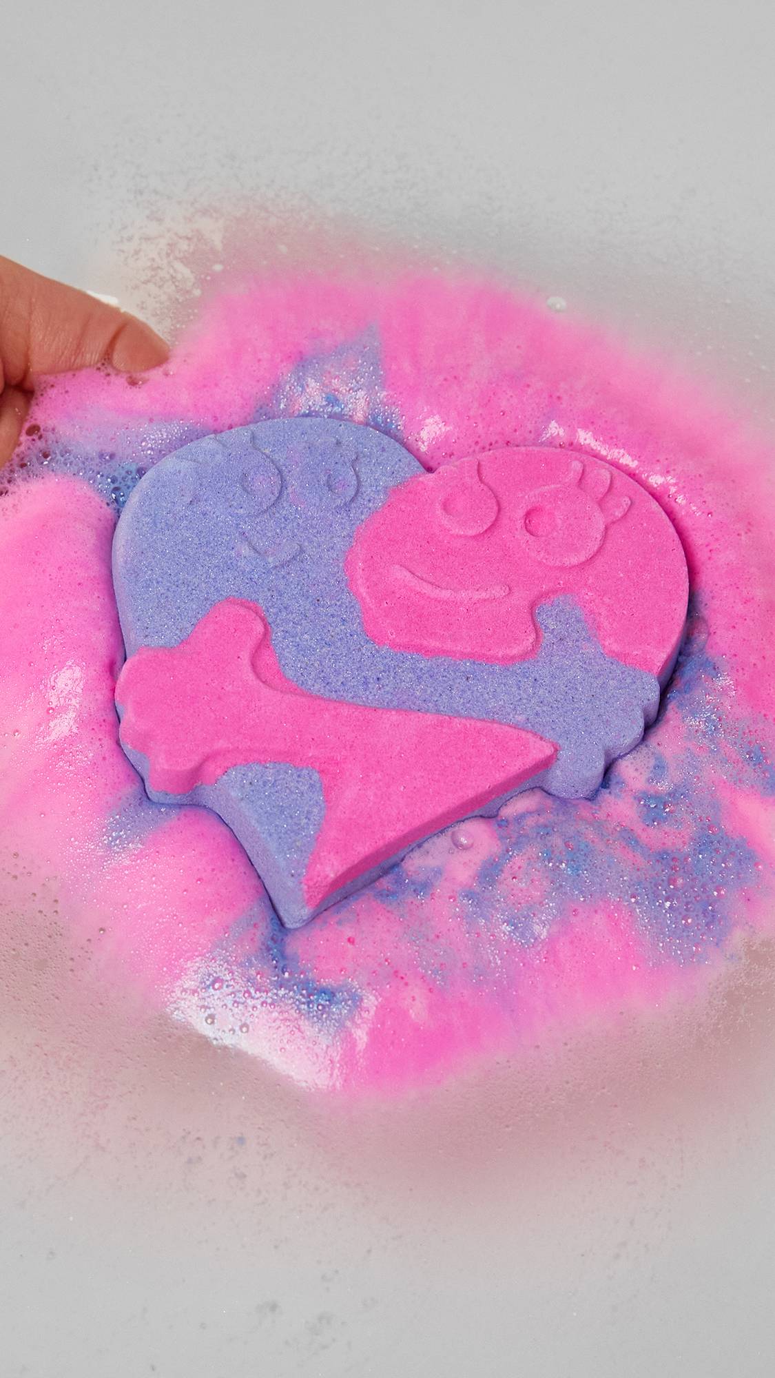 Image shows a close-up of the model gently placing the Big Squeeze bath bomb into the water as vivid purple and pink foam is expelled.