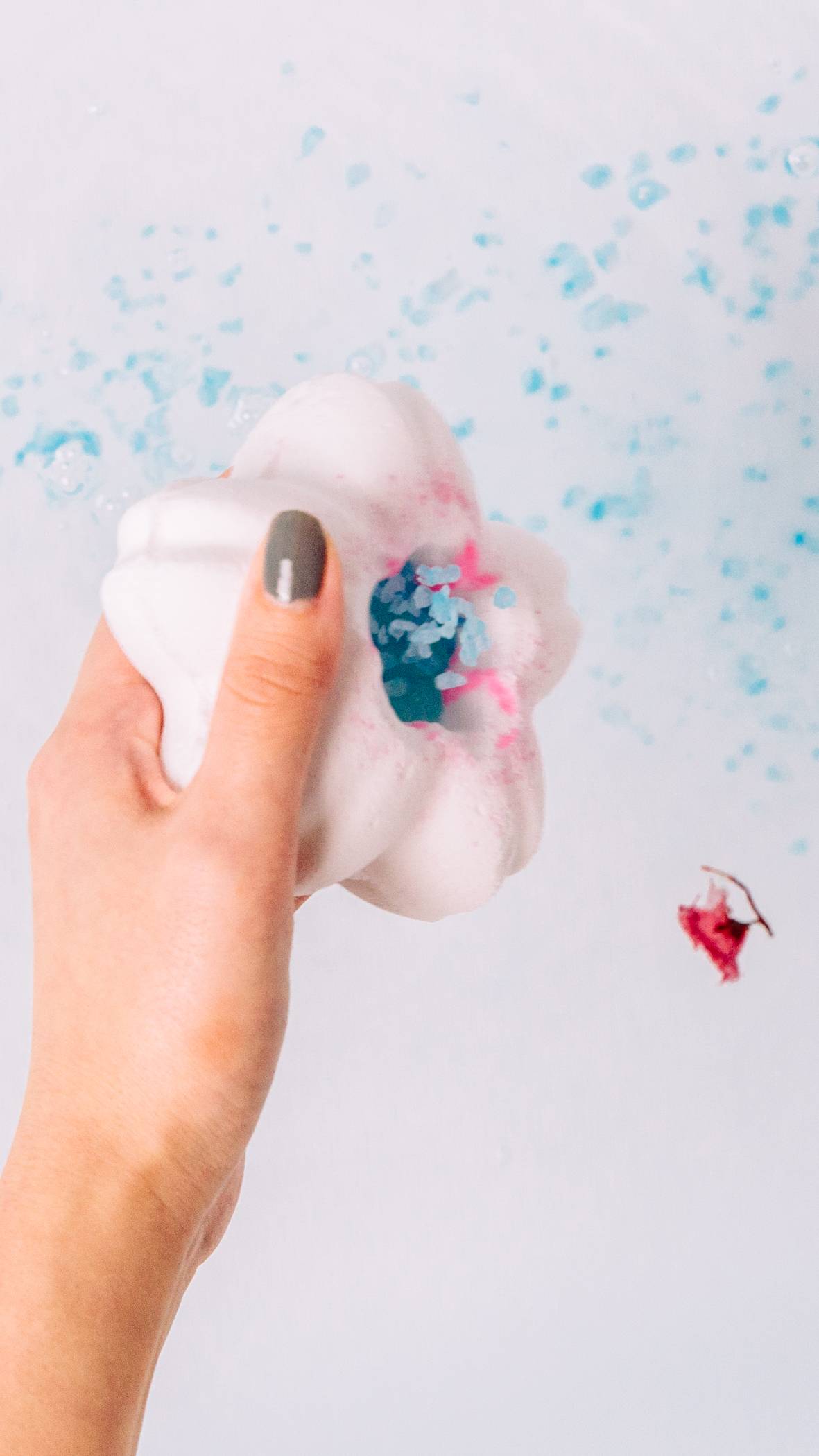 The image shows the model sprinkling blue bath salts from the white bath bomb flower part into the bath water below. 