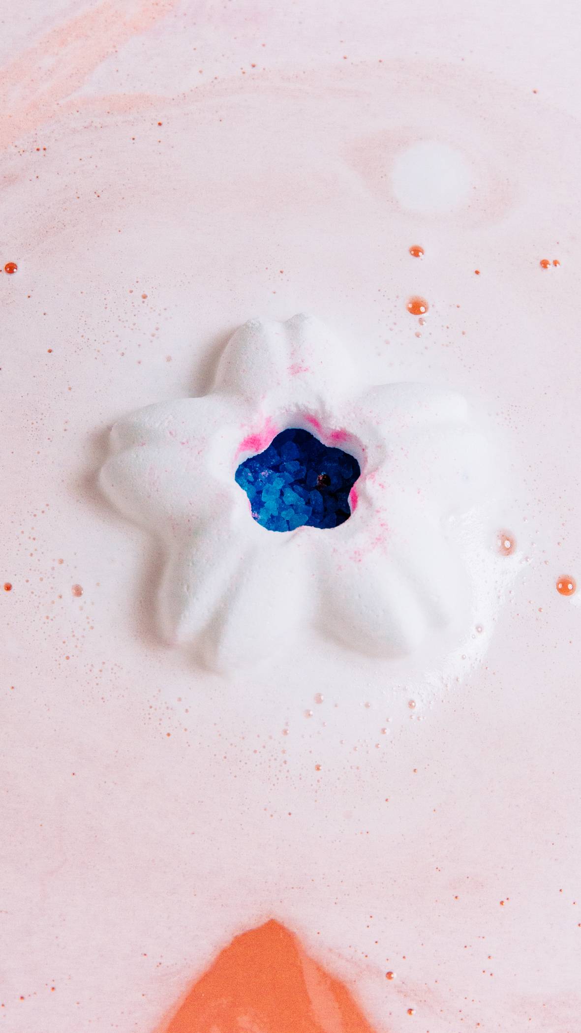 The pink lid of the bath bomb has been taken off and the main, hollow section of the bath bomb is sitting in the bath filled with blue salt crystals just visible inside. 