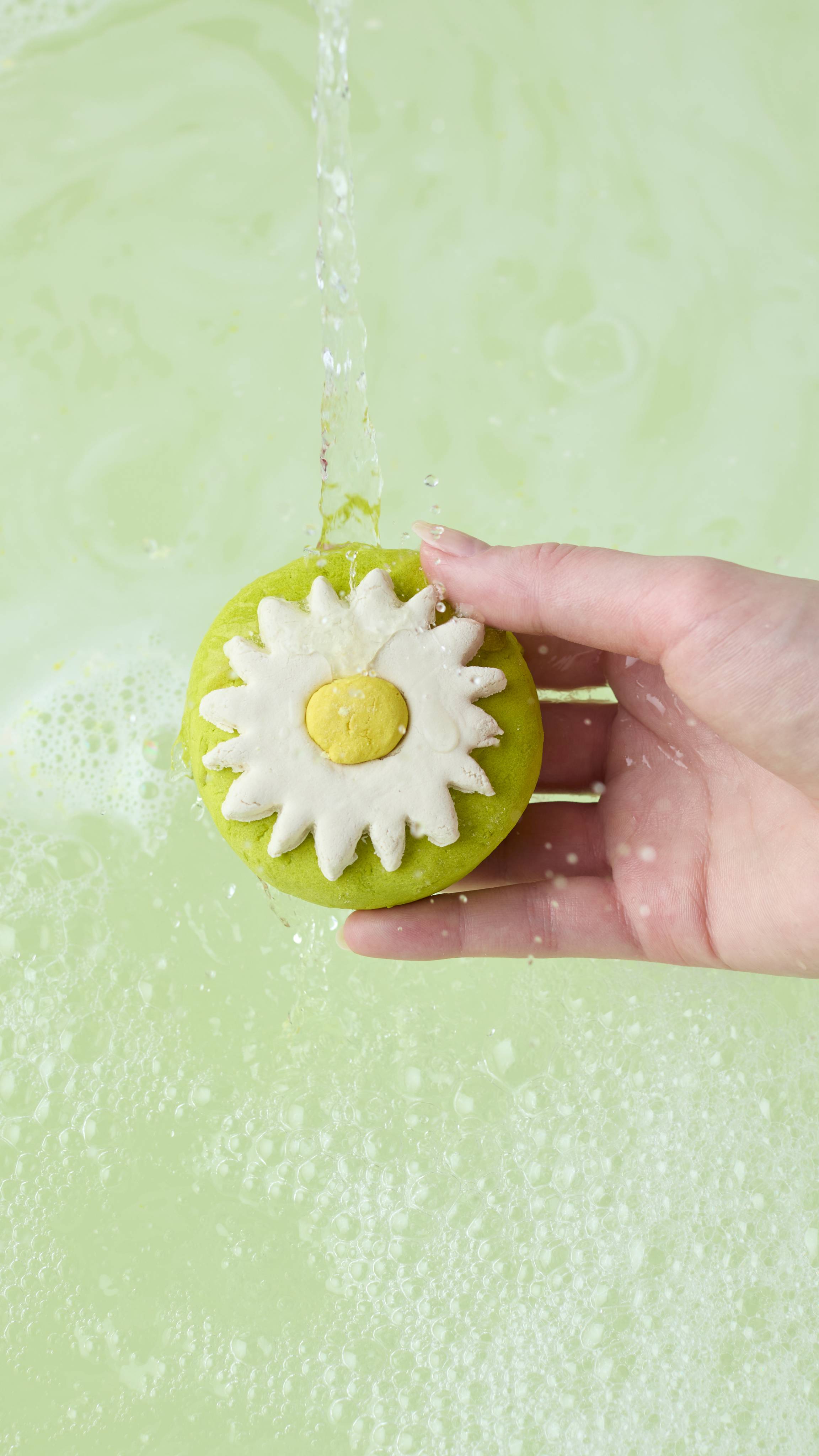 A hand holds the "Blooming Beautiful" bubble bar under a running tap, allowing the stream of water to hit the white flower and yellow center. The running water is generating bubbles in the pale green bathwater below.