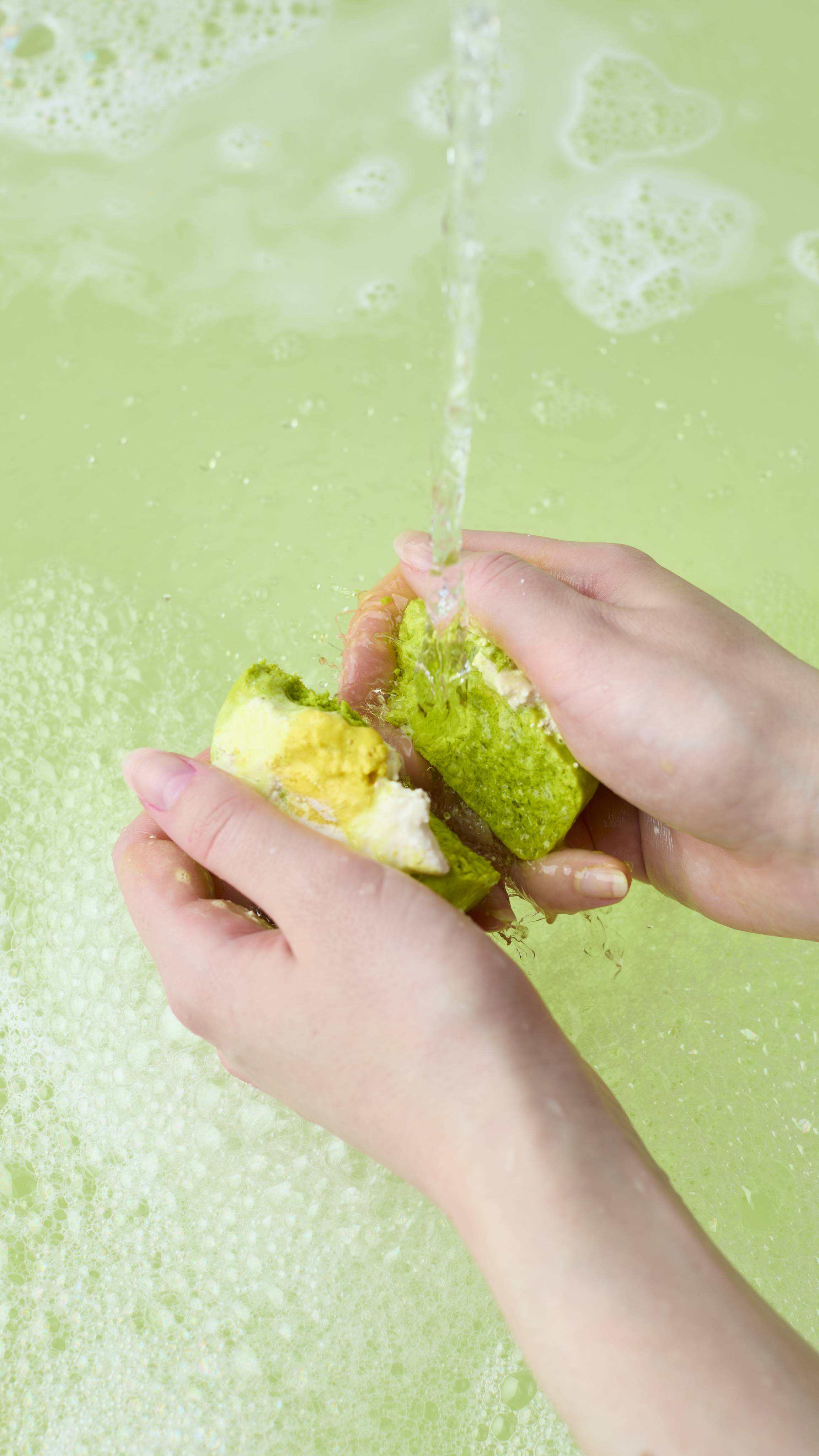The image shows two hands crumbling a green and white "Blooming Beautiful" bubble bar under a running tap in a bath. The product dissolves into the pale green water, creating a thick, foamy layer of white bubbles across the surface.