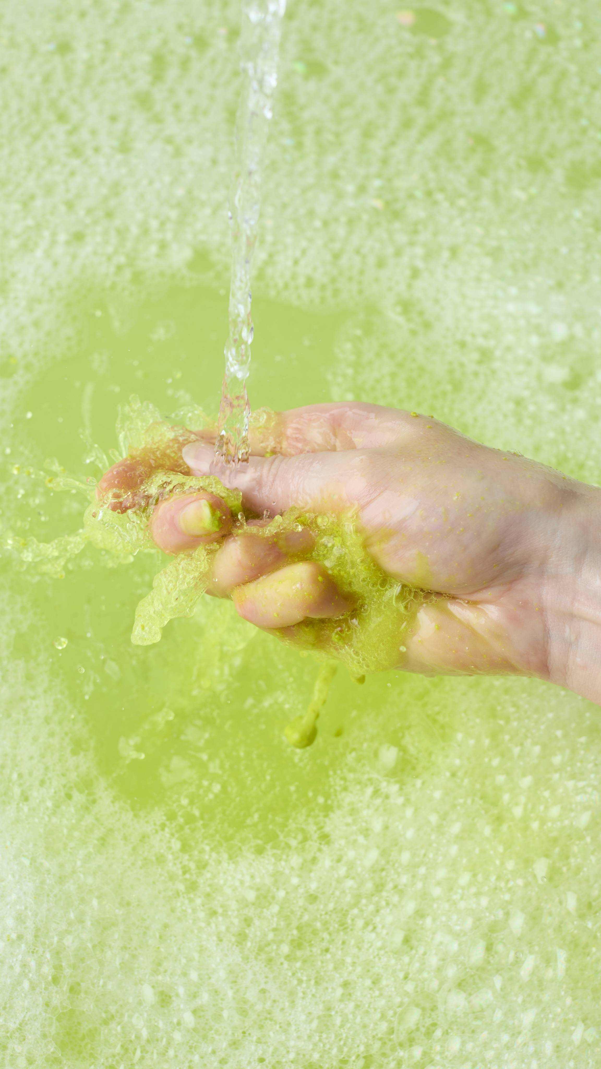 A hand holds the dissolving remnants of the green "Blooming Beautiful" bubble bar under a running tap. The water stream breaks down the product in the palm, turning the bathwater a vibrant lime green surrounded by soft white foam.