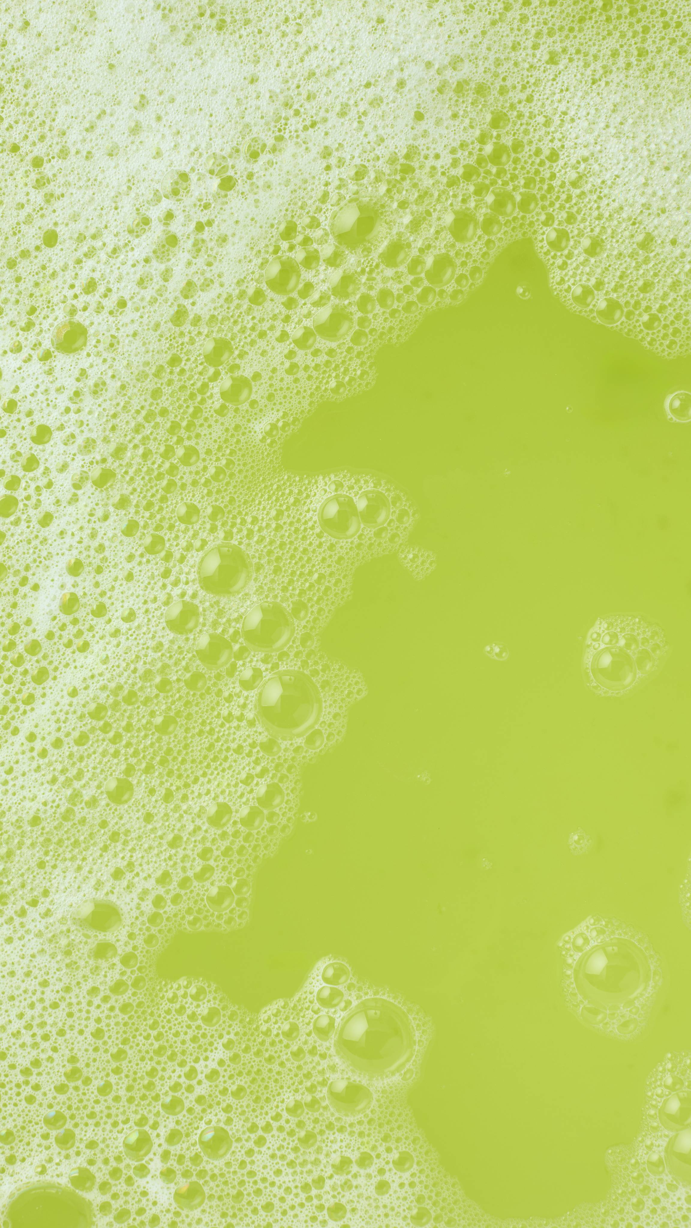 A top-down close-up shows the surface of the bathwater, which has turned a vibrant lime green. Patches of white frothy bubbles float on the colorful water, highlighting the result of using the bubble bar.