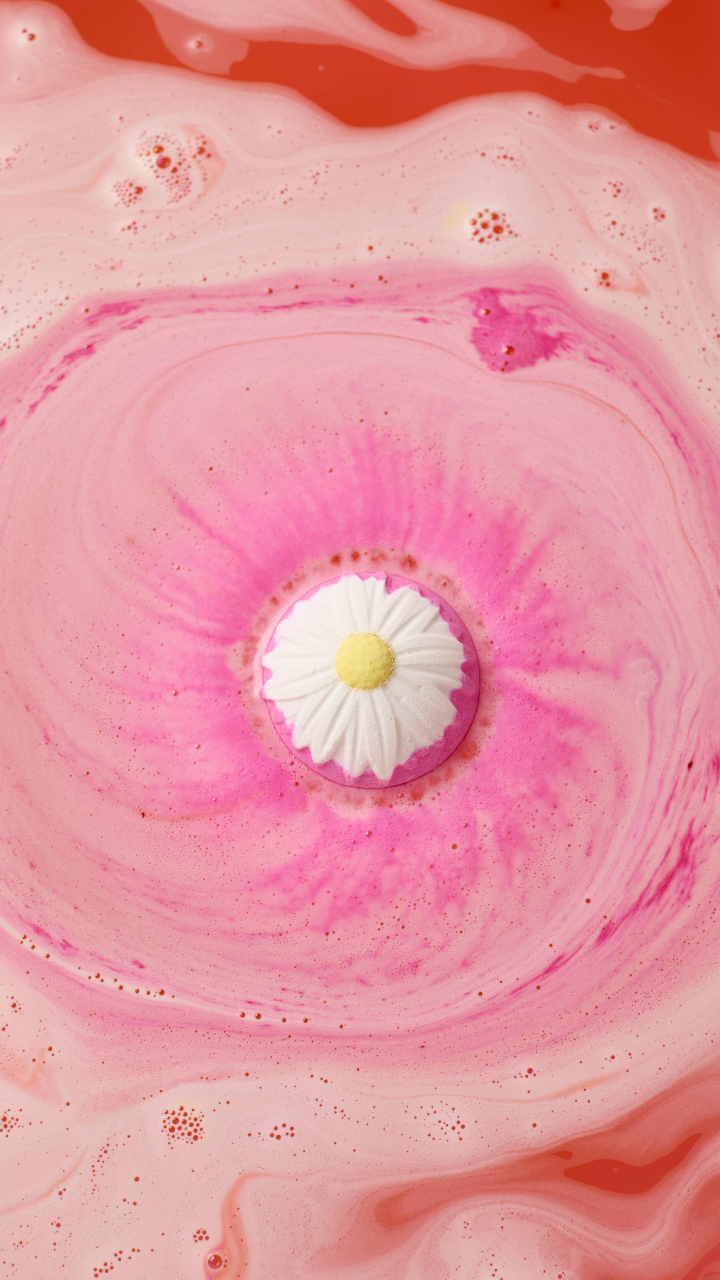 An aerial view of the "Blooming Beautiful Orange Blossom" pink bath bomb, with a white and yellow daisy pattern dissolving in water. It creates a swirling, vibrant pink foam that expands across the surface of the warm, peach-coloured bathwater.