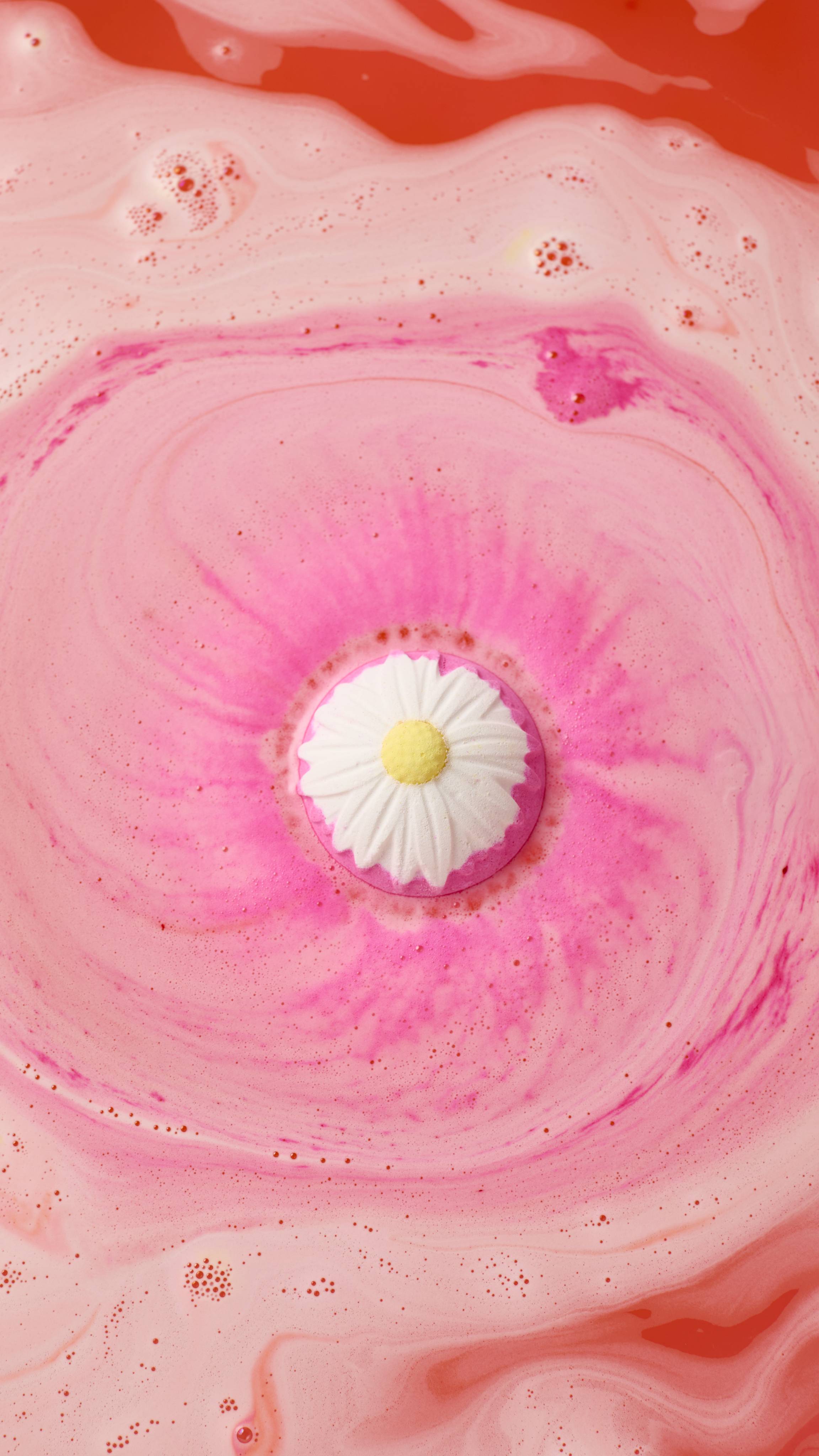 An aerial view of the "Blooming Beautiful Orange Blossom" pink bath bomb, with a white and yellow daisy pattern dissolving in water. It creates a swirling, vibrant pink foam that expands across the surface of the warm, peach-coloured bathwater.