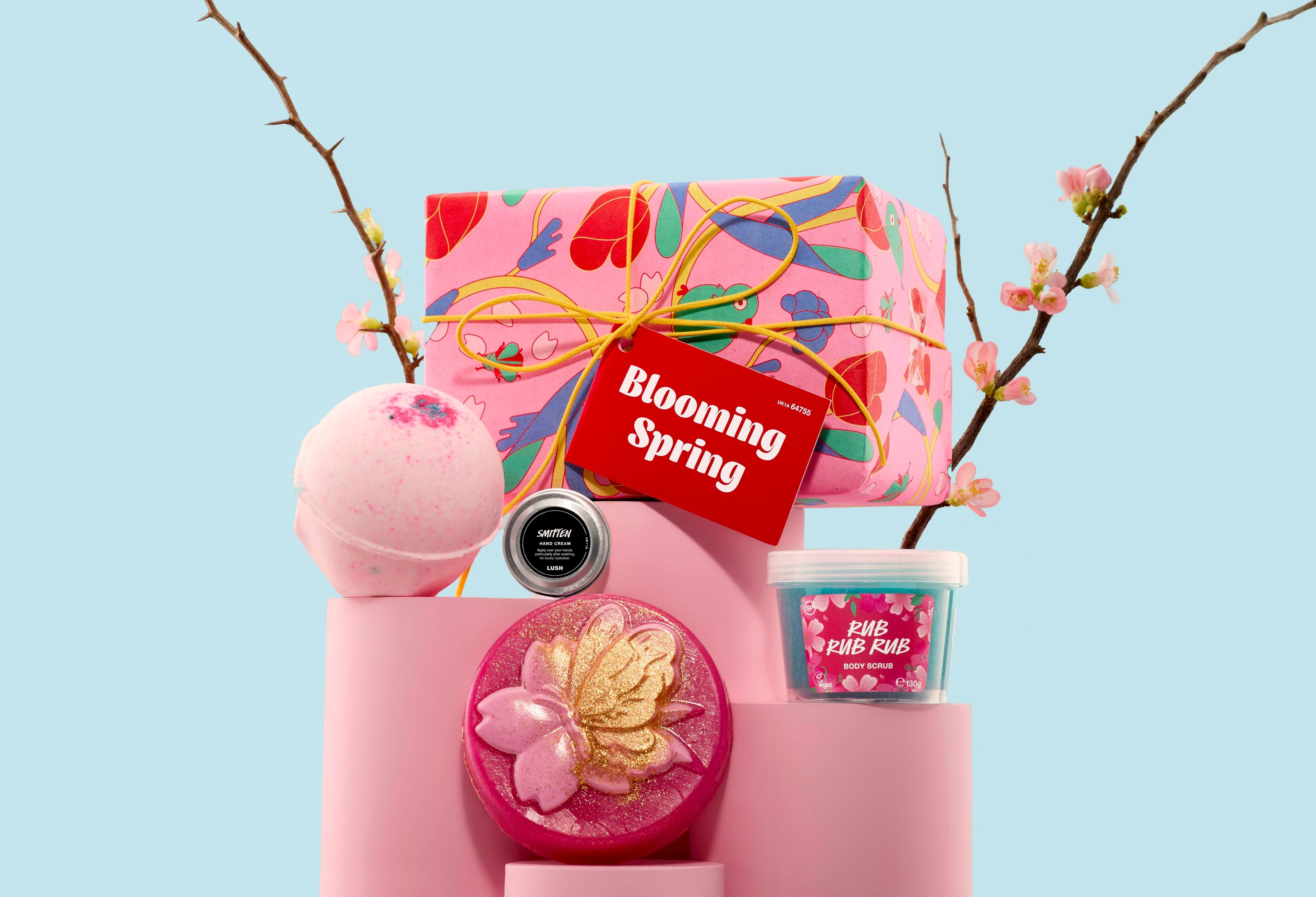 The image shows a gift set featuring a wrapped pink box with a "Blooming Spring" tag, a bath bomb, a small tin, a circular soap, and a pot of body scrub. All items are arranged on pink podiums with blossom branches against a blue backdrop.