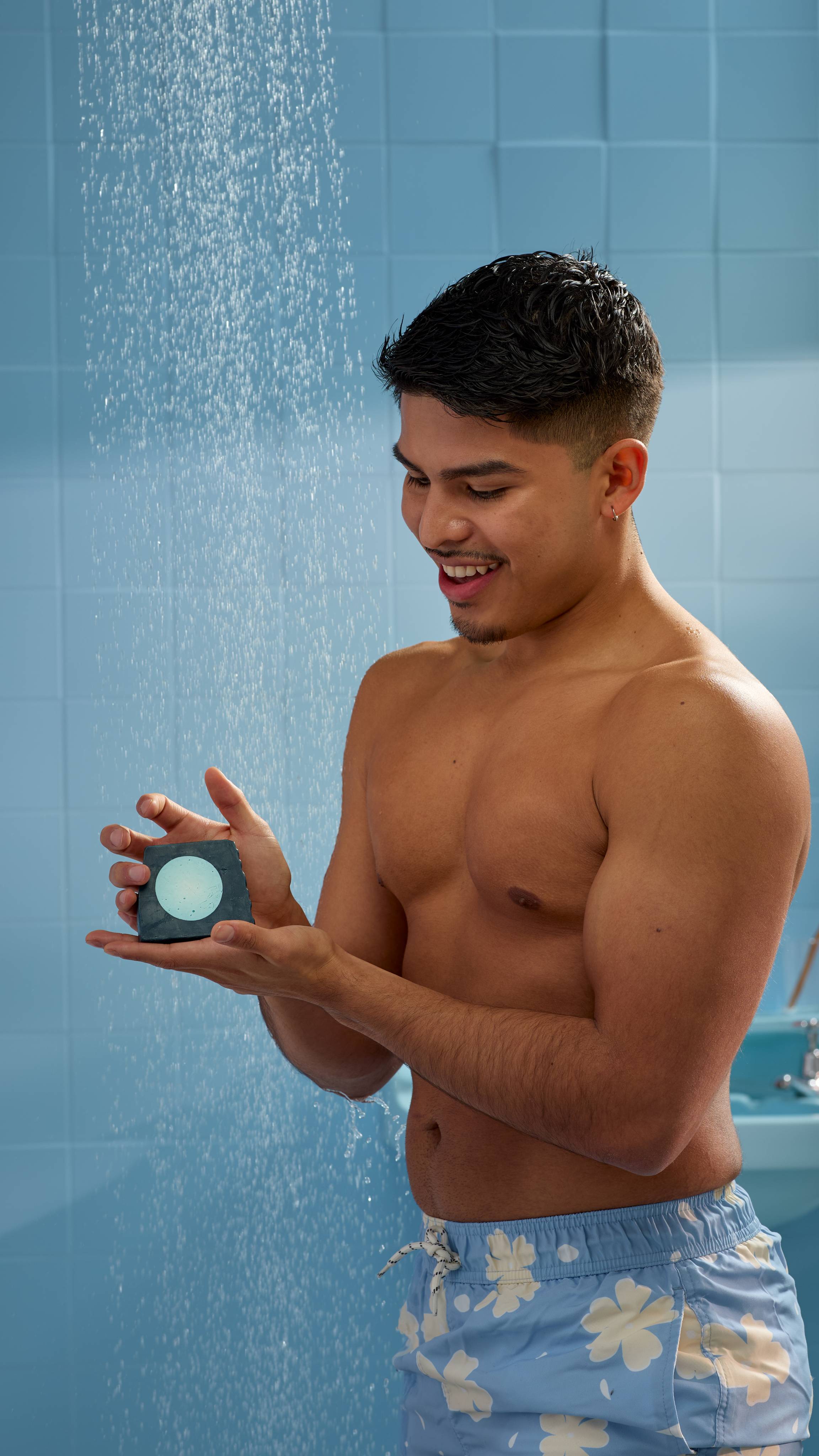 The model is in the shower in a blue, tiled bathroom. They are in blue and white shorts holding the Blue Moon soap with both hands and water falls behind them. 