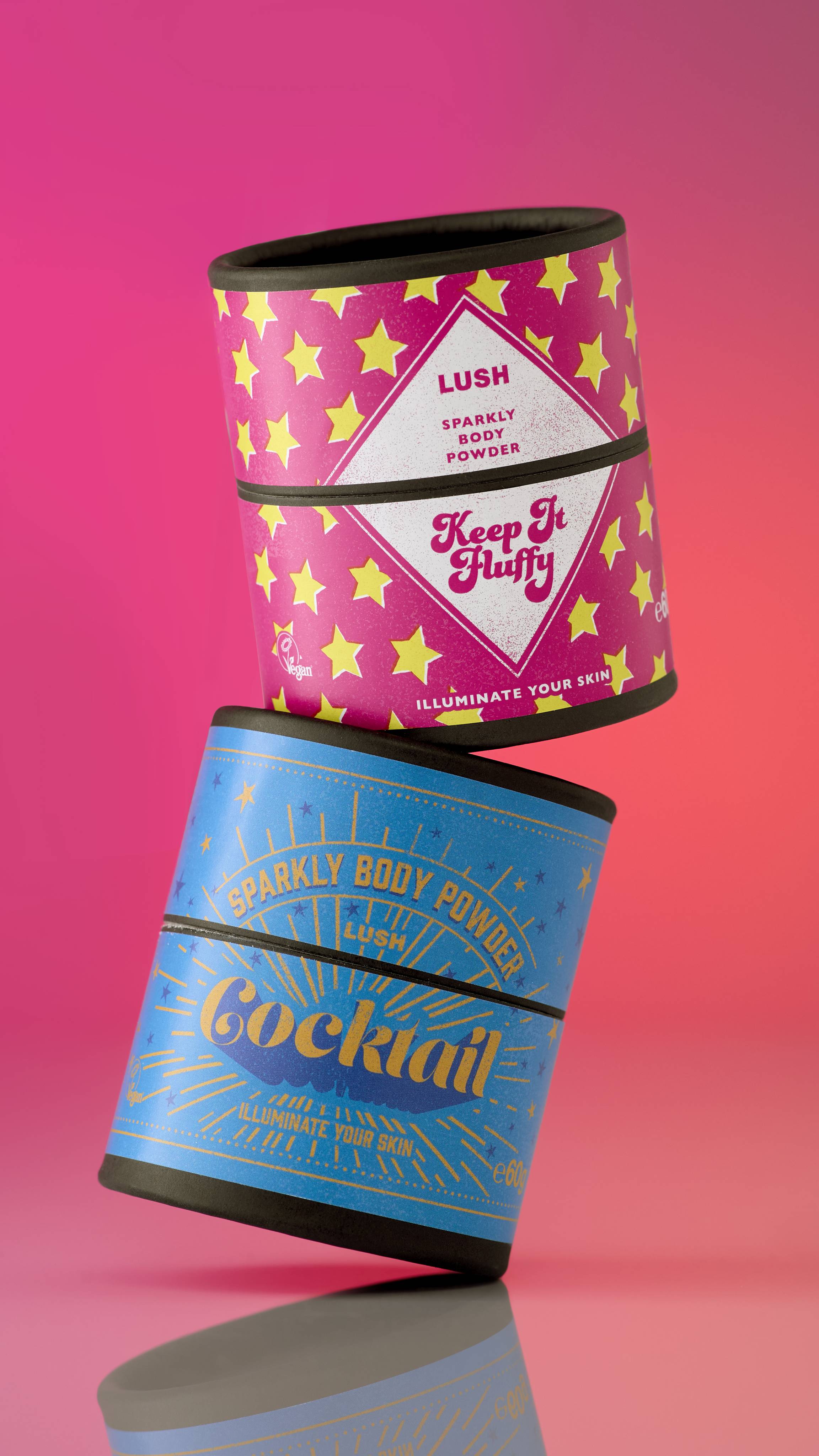 Two Lush sparkly body powder shakers are stacked on an orange-pink background. The top "Keep It Fluffy" container is magenta with stars. The bottom "Cocktail" container is blue with stars. They are slightly angled and appear to be floating.