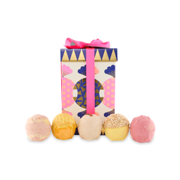 Bon Bon. A rectangular-shaped box with a lid sits upright. It is a cream colour with vibrant, wrapped sweet-shaped designs around it with gold detail. It is tied with a pink ribbon as five Lush bath bombs sit in front of it. 