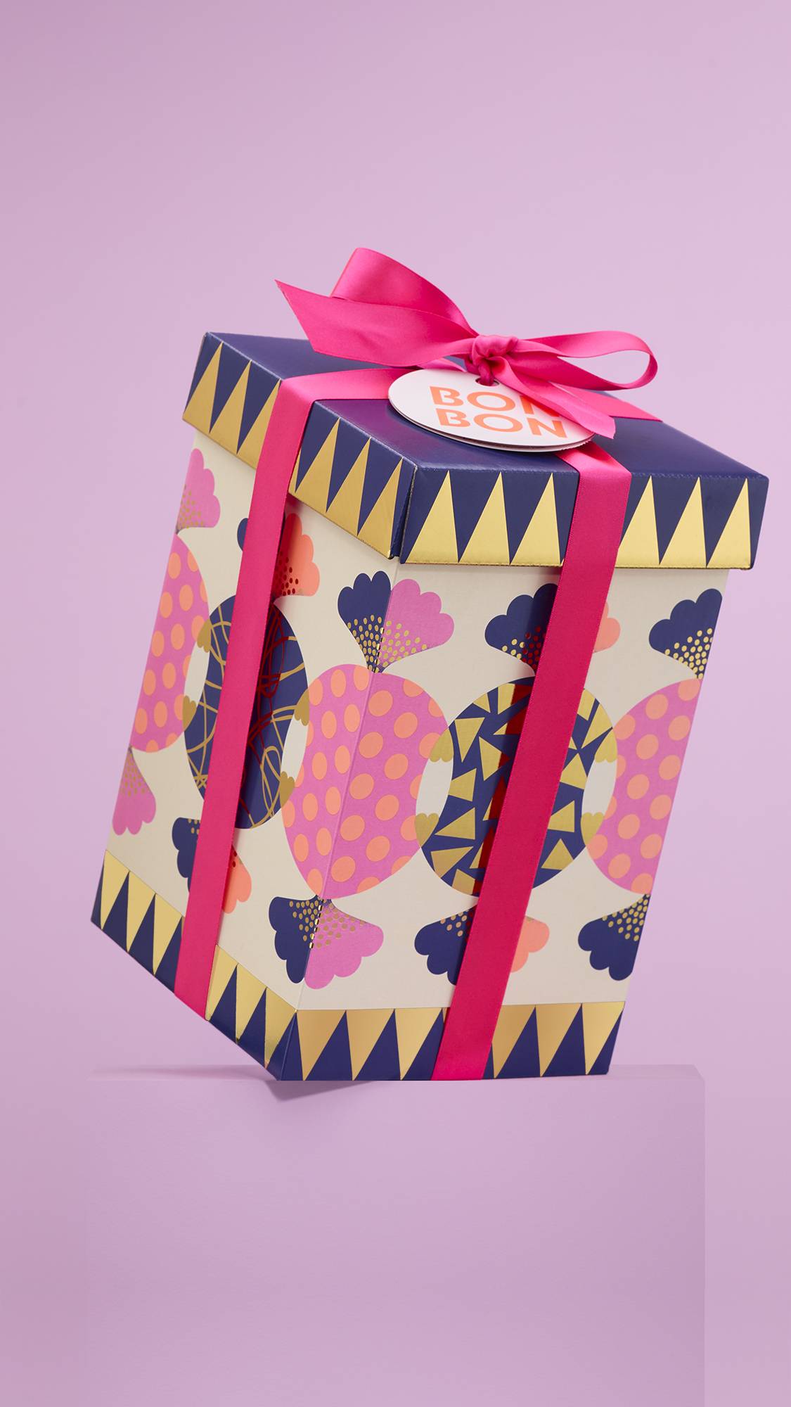 The image shows the large, sweet-designed "Bon Bon" gift box, tied with purple ribbon and a white gift tag, sitting on a pale-pink platform against a matching pink background. 
