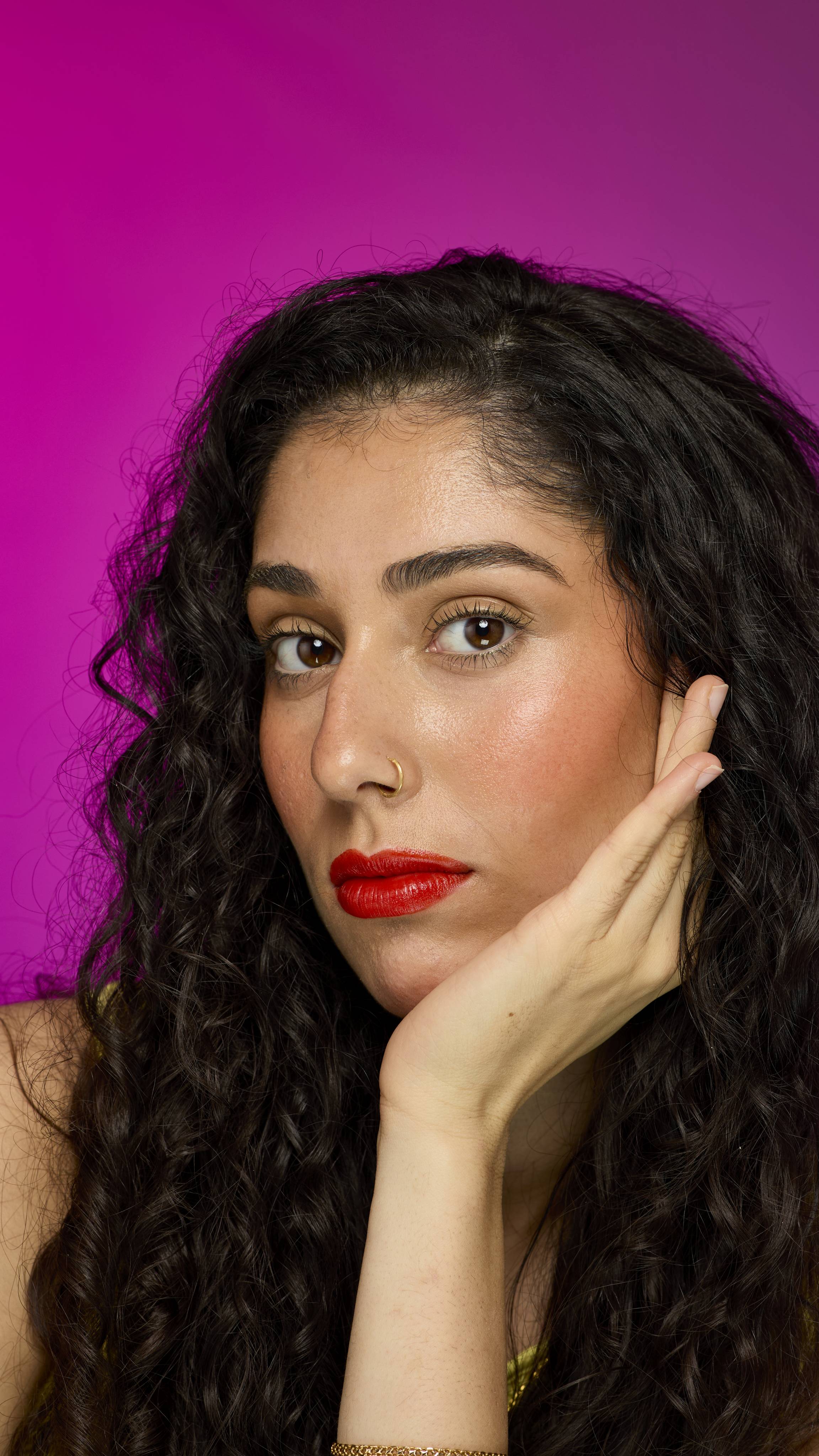 A model with long, dark hair is shown from the neck up. They are on a vibrant pink background, showing off the vibrant, ruby-red Bonfire Night lipstick as they rest their chin on their hand.
