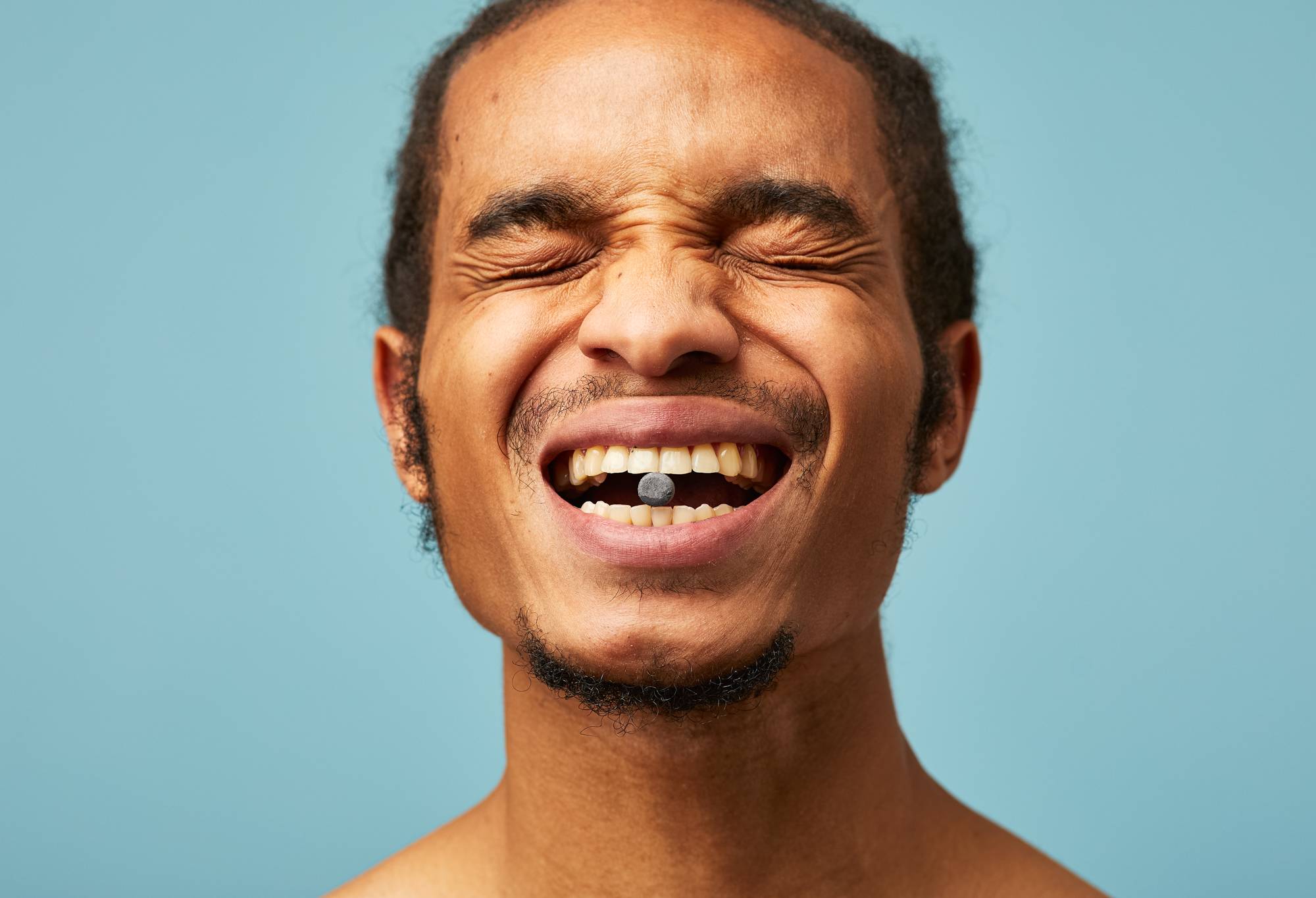 A person shows off their teeth and holds a small black Boom! toothpaste tab between their front teeth.