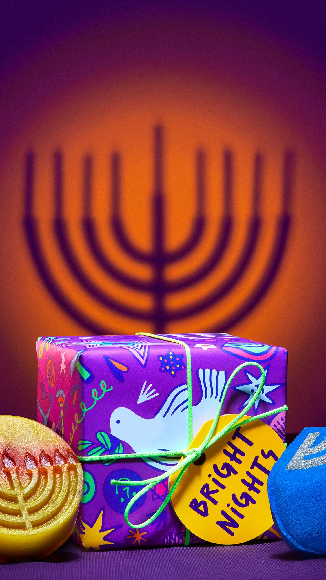 The image shows the Bright Nights gift box set with the two included Hanukkah-themed products beside it. The background is a deep purple and orange with the shadow of a Menorah.