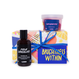 A small Brightness Within Gift set sits on a light grey background. It includes Posh Chocolate Shower Gel and Sufganiyah Sugar Scrub, placed on a colorful, patterned box tied with a blue ribbon and a bright blue tag.