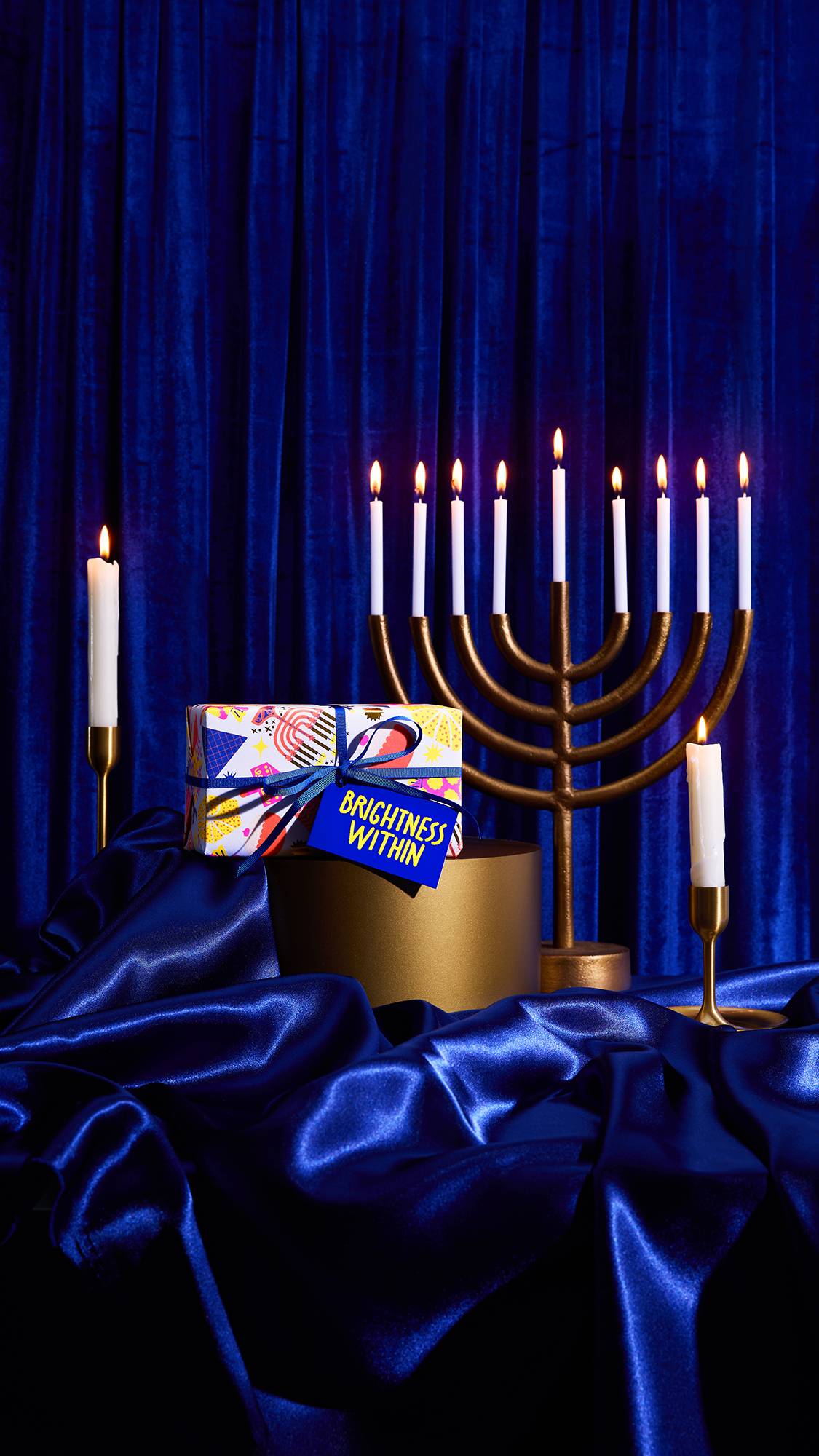 A dramatically lit Brightness Within Gift box sits on a gold cylinder next to a gold menorah with eight lit candles. Two single lit candles are also visible against a rich royal blue velvet and satin backdrop.