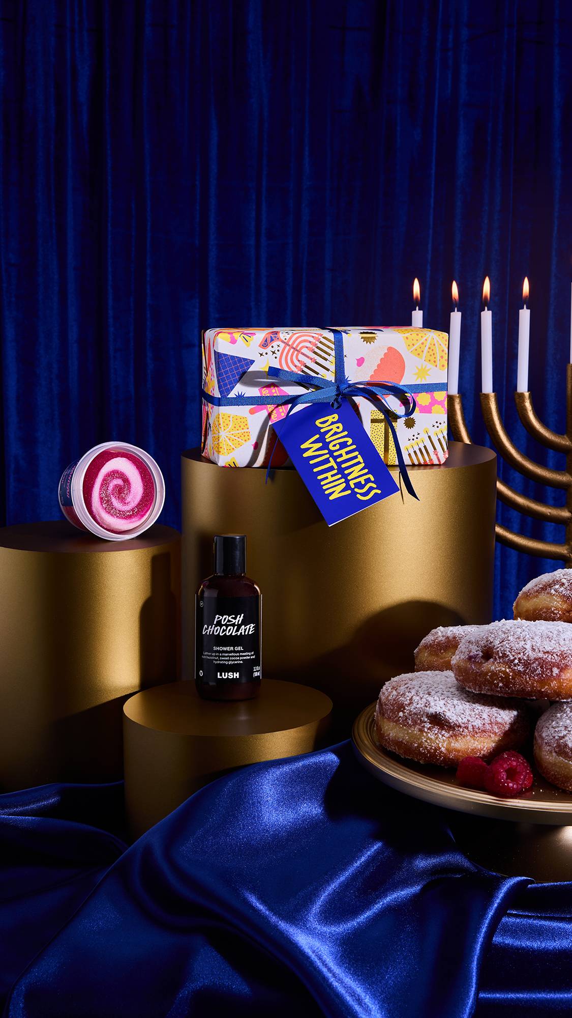 A scene features the Brightness Within Gift box, Posh Chocolate Shower Gel, and swirled Sufganiyah Sugar Scrub displayed on gold props. A menorah with four lit candles and donuts are visible against a royal blue velvet backdrop.