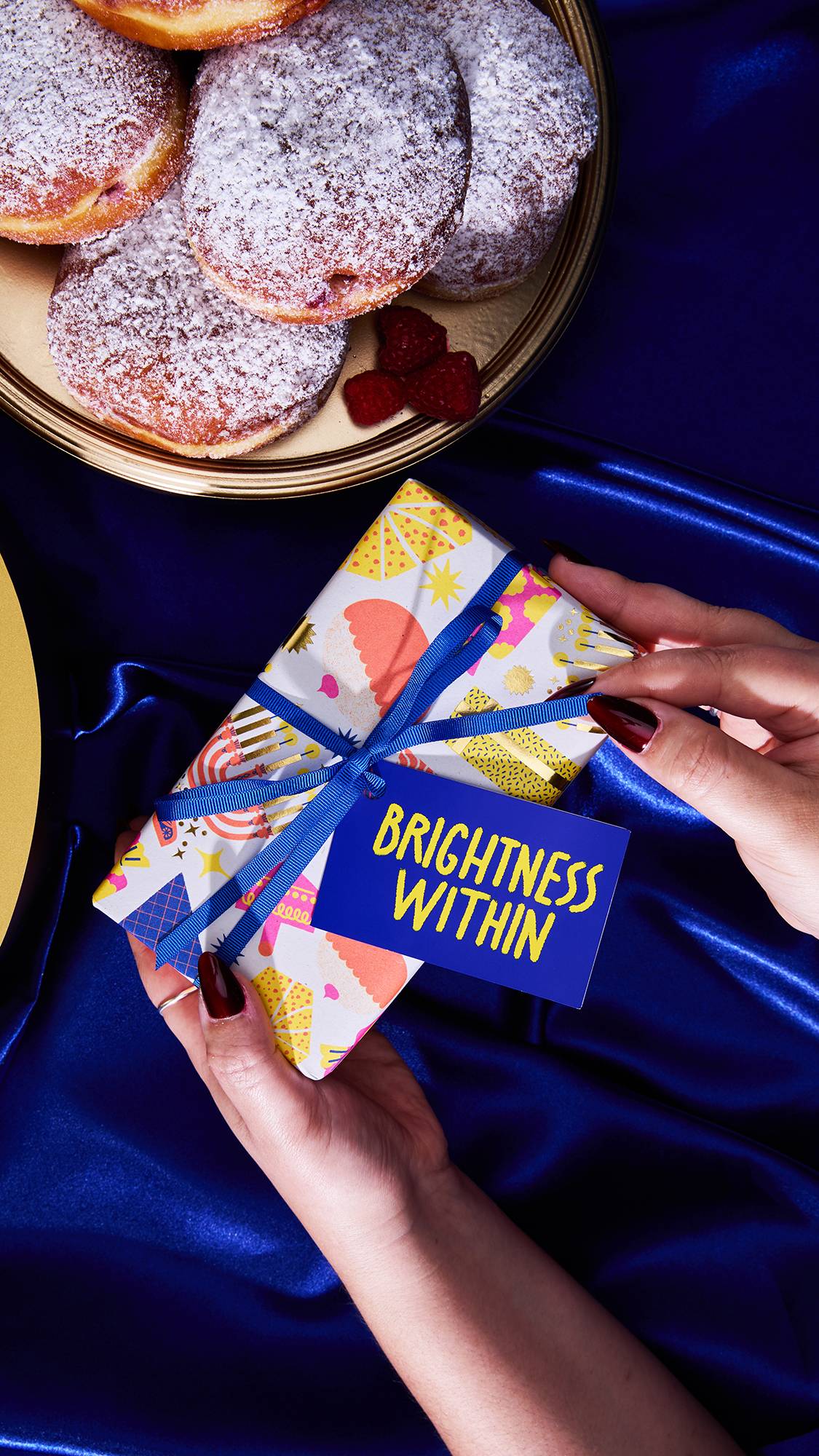 Two hands with dark nail polish hold the brightly patterned Brightness Within Gift box, tied with a blue ribbon and tag, over deep royal blue satin fabric. A platter of powdered sugar donuts topped with raspberries is visible in the background.