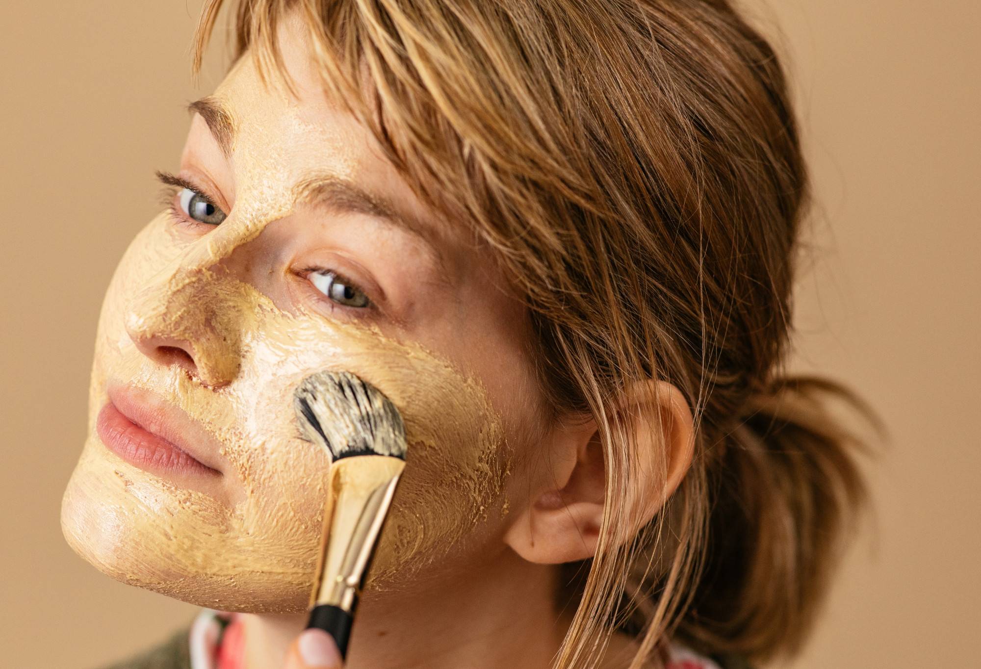 Smooth, light gold coloured Brush Strokes face mask has been applied to the face with a make up brush, finishing on the cheeks.