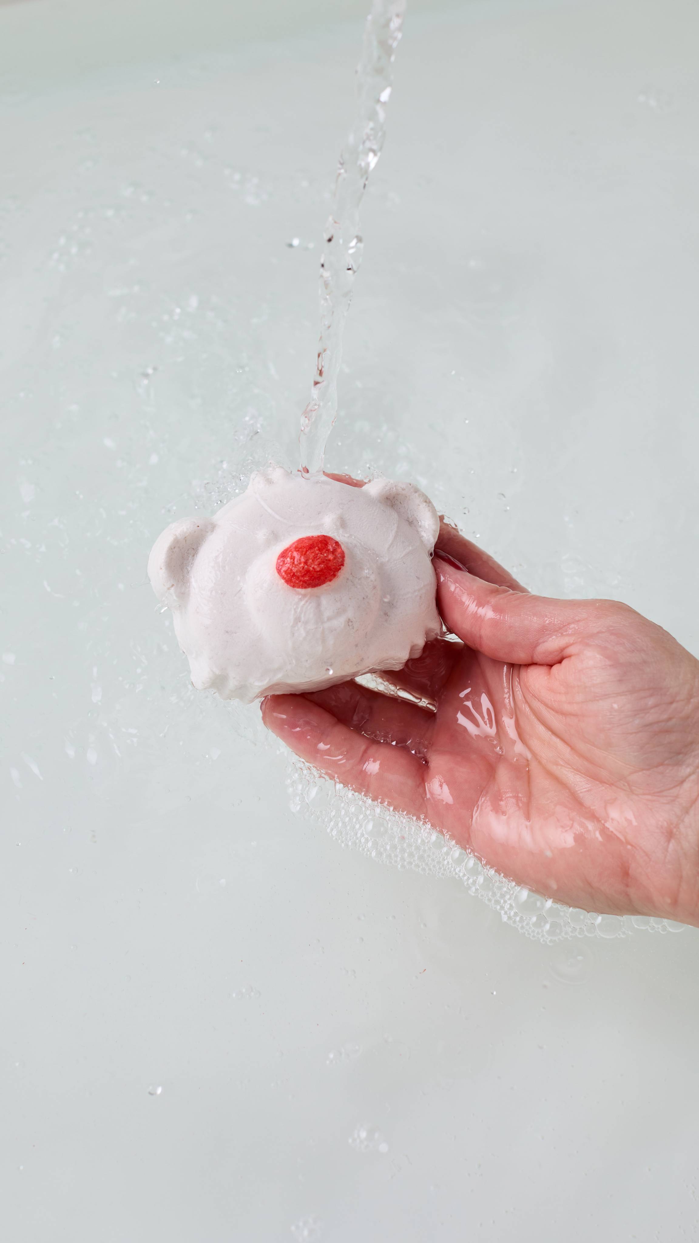 A close-up of the model's hand reaching in from the right while holding the Lush Christmas Bubble Bear bubble bar under running water. Delicate white bubbles form on the surface of the bath water below. 