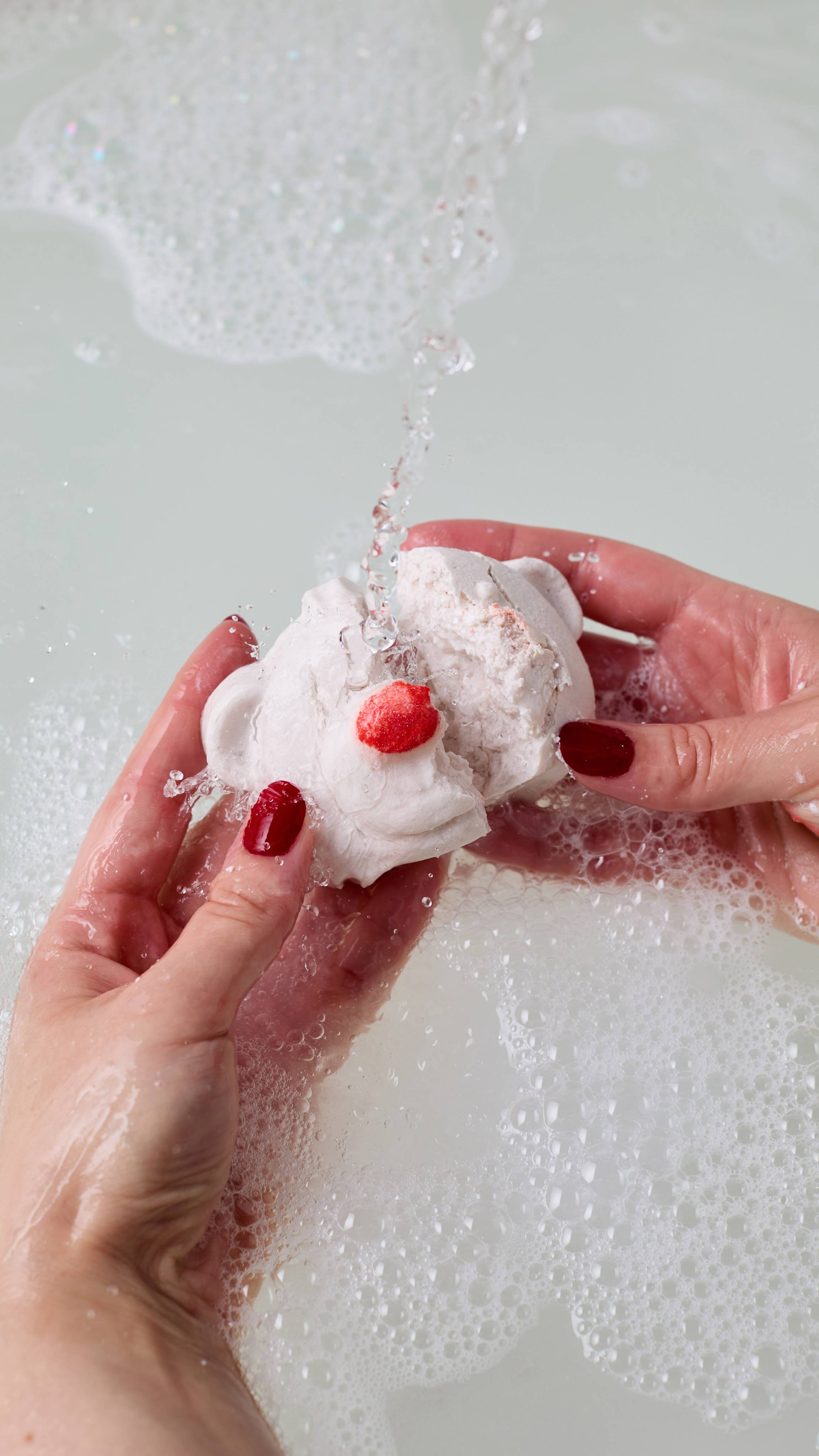 The image shows the model's using both hands to break the white Bubble Bear bubble bar under running water.  There is a thick layer of bubbles on the bath made by the product. 
