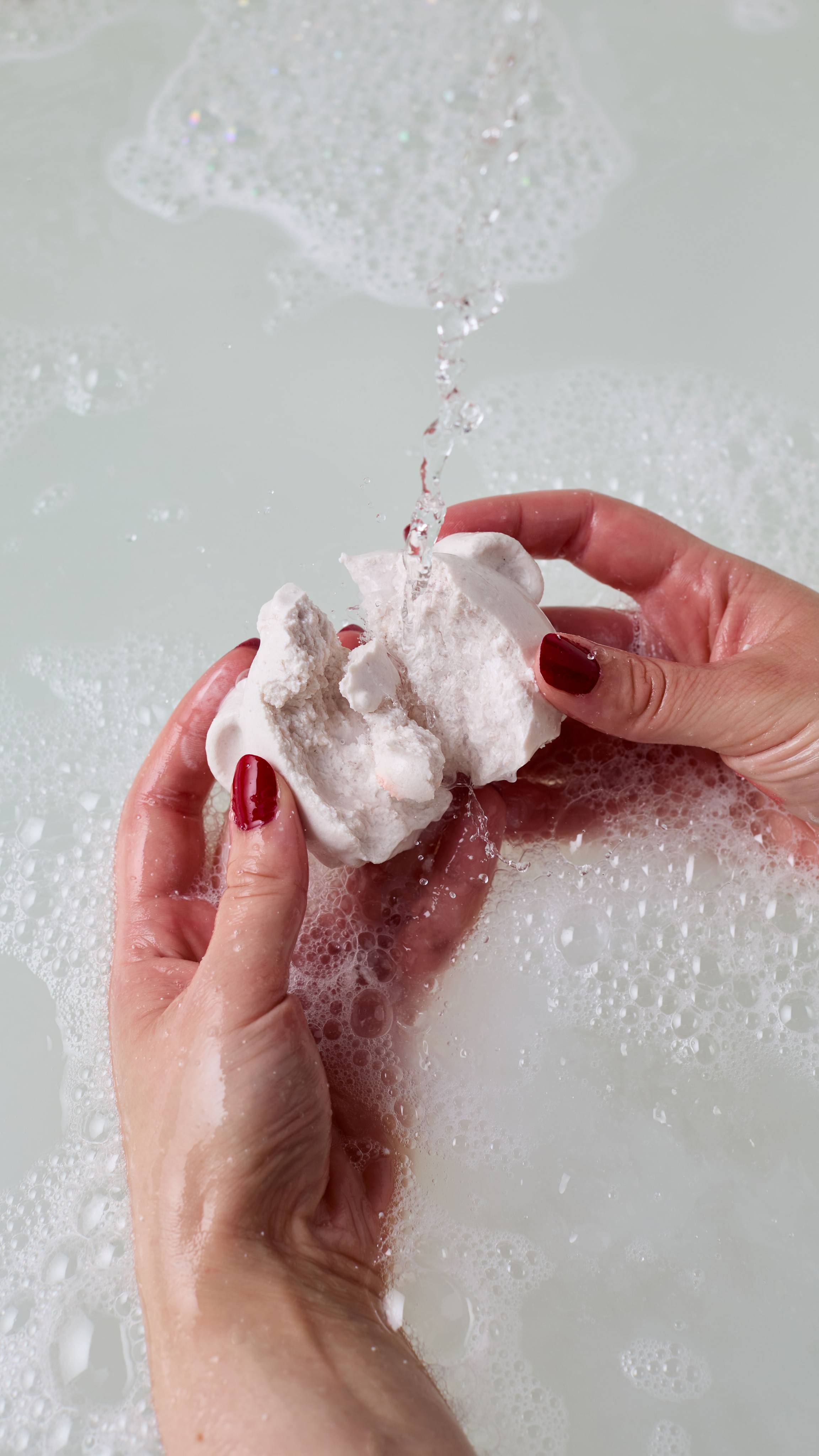 The image shows the model's using both hands to break the white Bubble Bear bubble bar under running water.  There is a thick layer of bubbles on the bath made by the product. 