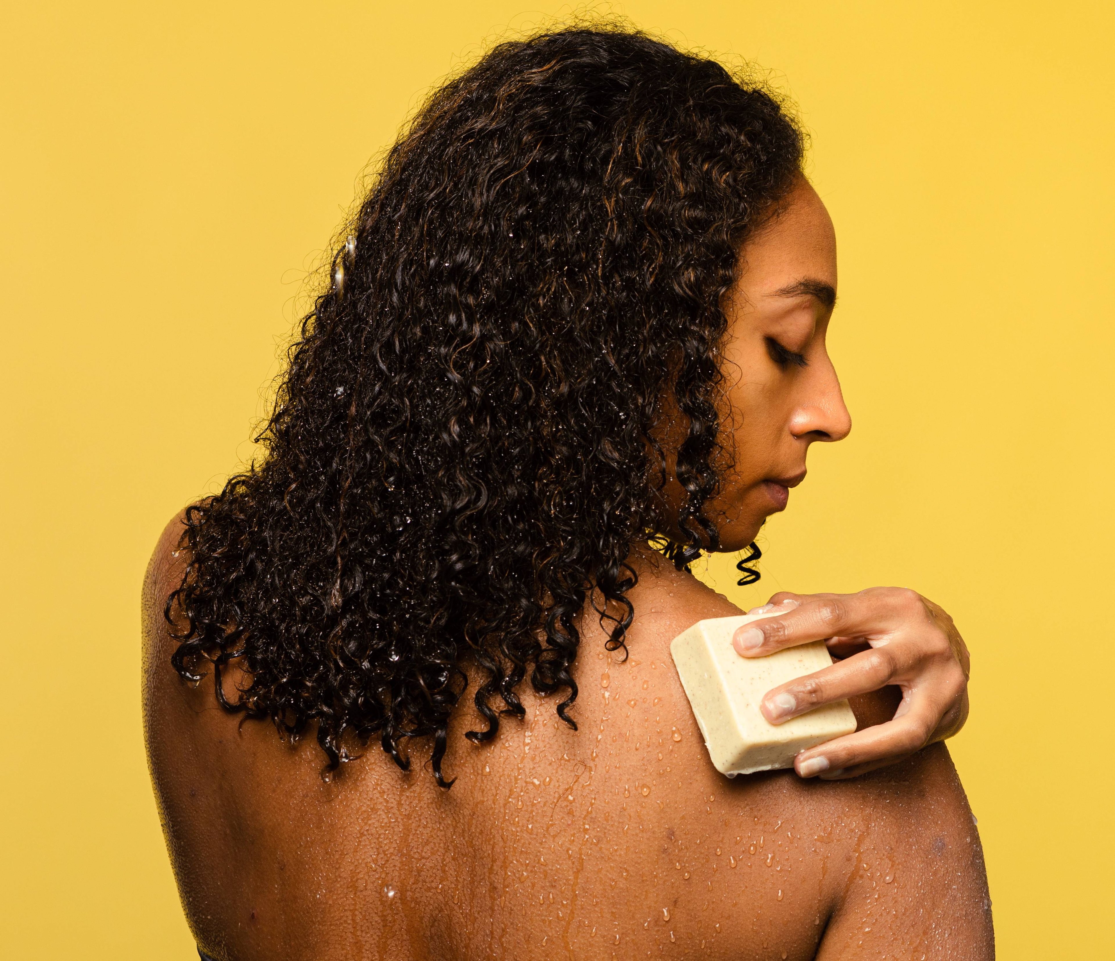 A person with long, curly hair holds Buffy, a cream coloured, square shaped solid body butter, on their water beaded shoulder.