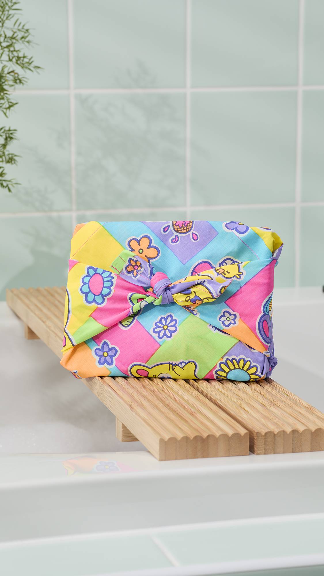 The image shows the colourful "Bunnies Picnic" knot wrap, tied in a traditional Japanese knot, resting on a slatted wooden bathtub tray. The wrap features colourful flowers and characters in bright yellow, pink, and blue. 