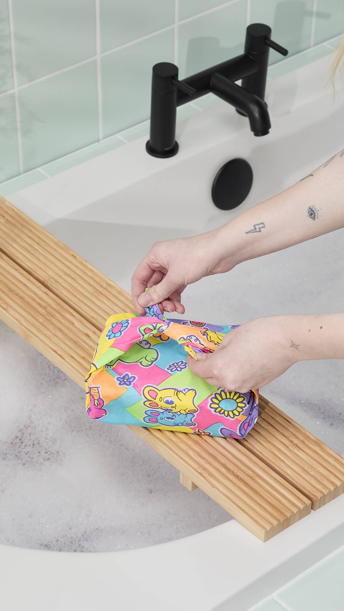A model's tattooed arms reach over a white bathtub to untie a "Bunnies Picnic" knot wrap tied neatly around a gift. The parcel sits on a slatted wooden bath tray above soapy water, with a modern black tap and mint tiles in the background.