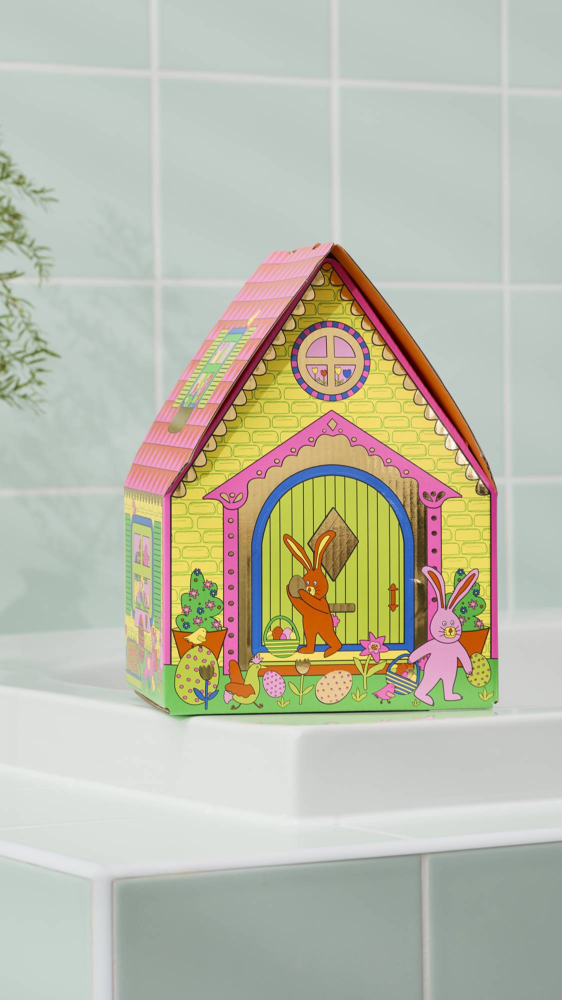 A house-shaped "Bunny Cottage" Easter gift tin sits on the white edge of a bathtub. The colourful tin features illustrations of bunnies, Easter eggs, and chickens against a yellow brick background. All set against pale green bathroom tiles.