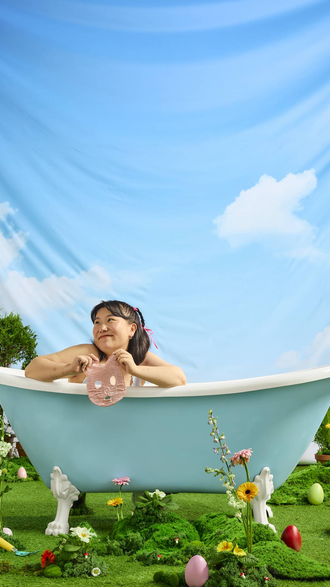 The model is sat in the outdoor, roll-top bath under blue skies as they hold the shimmery Bunny face mask. Grass and fresh spring flowers cover the floor. 