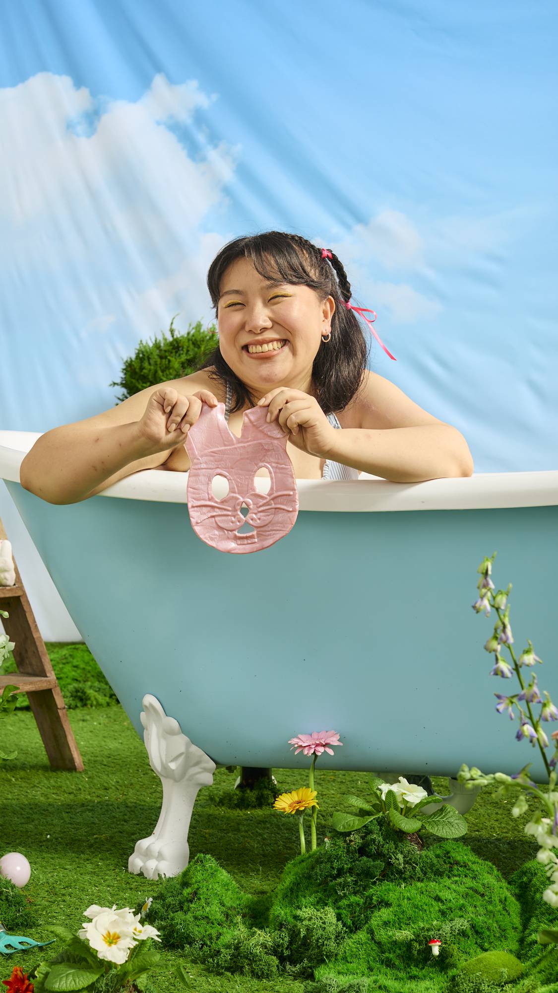 The model is sat in the outdoor, roll-top bath under blue skies are they hold the shimmery Bunny face mask. Grass and fresh spring flowers cover the floor. 