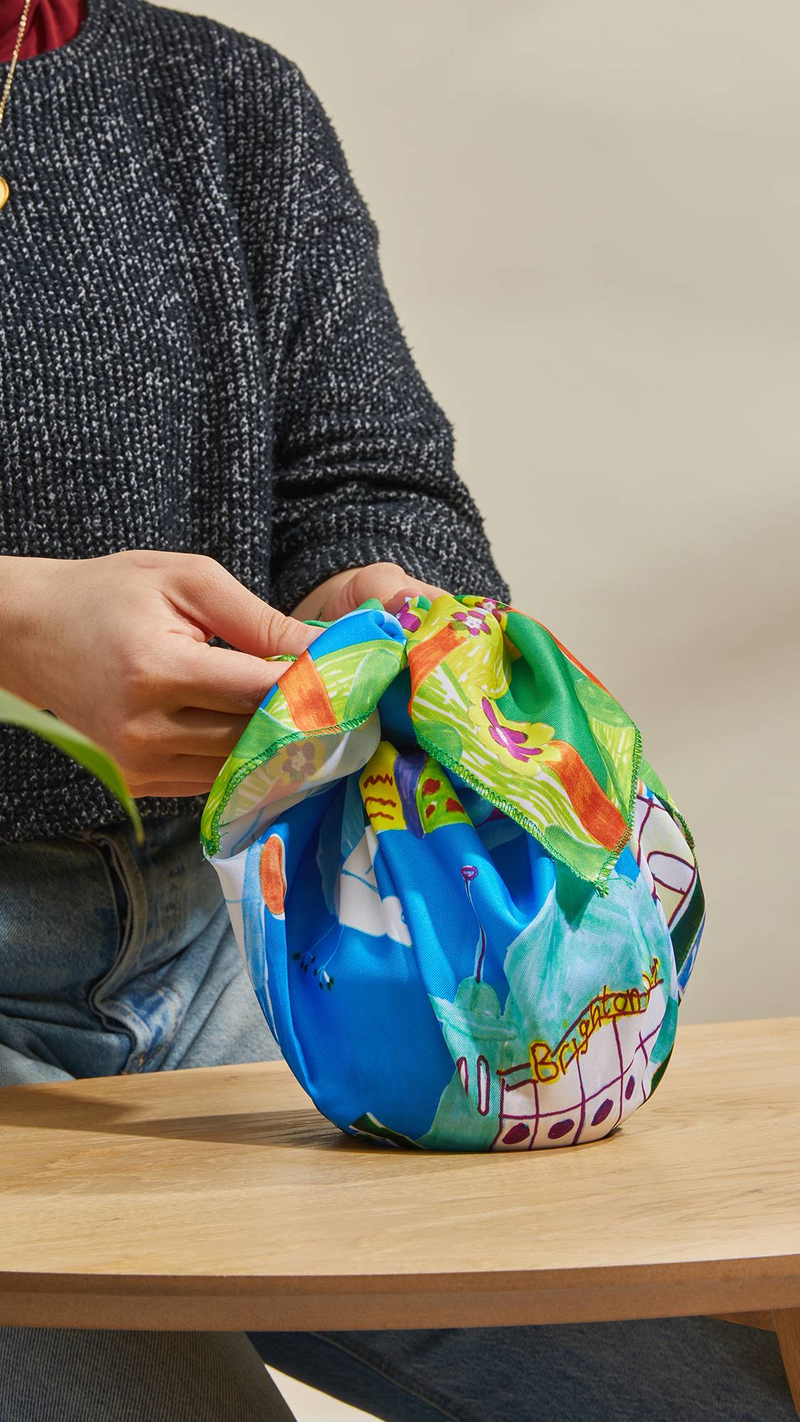 The image shows the model's hands as they stand at a counter and neatly tie the By The Seasing knot wrap around a gift.