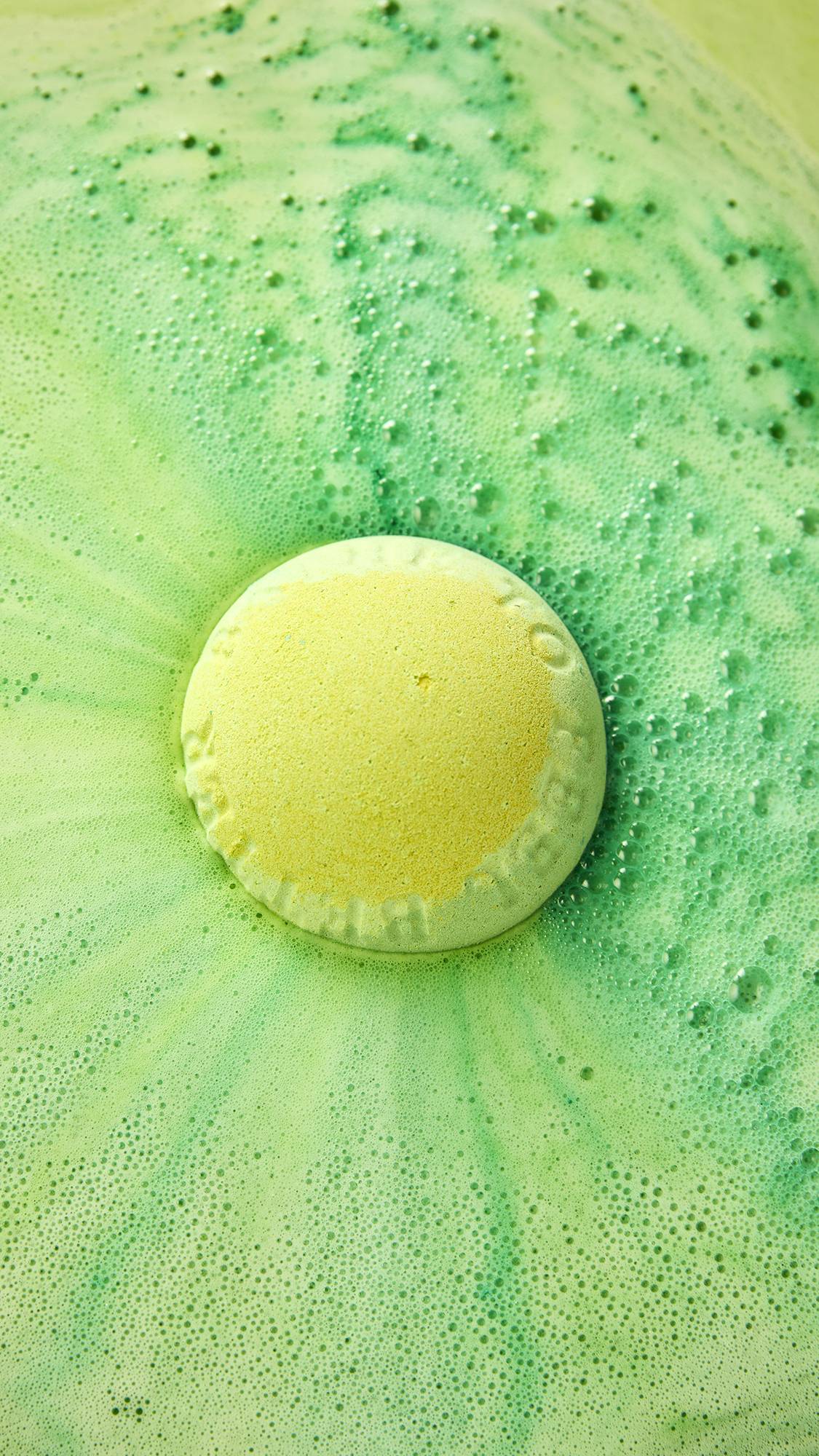 A yellow and green LUSH "Byegraine" bath bomb dissolves in bright green bathwater, floating in the centre as it produces a radiating pool of fine white foam and tiny fizzing bubbles.