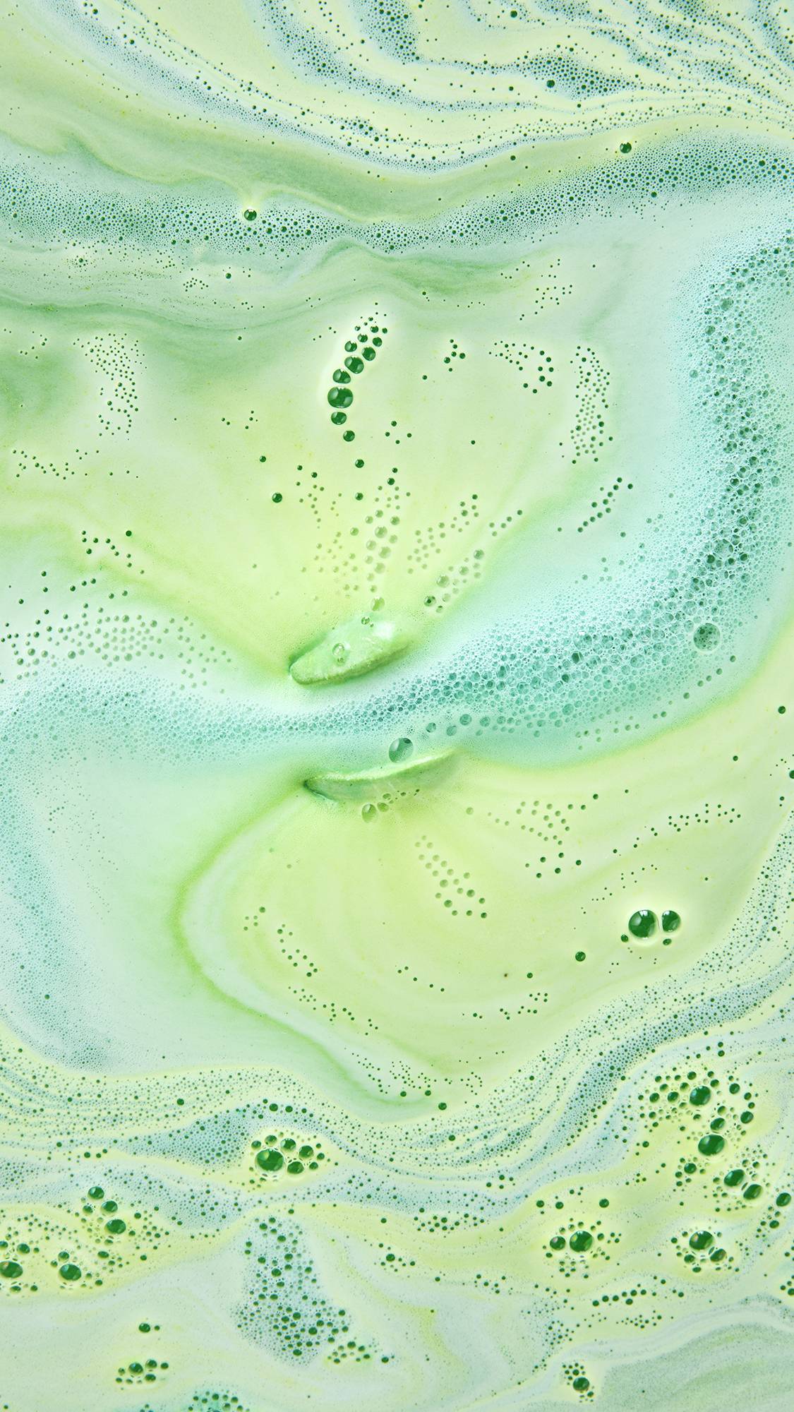 An aerial close-up of bathwater with swirling marble patterns of pale lime-green and light mint-blue foam, dotted with clusters of fine bubbles across the surface.