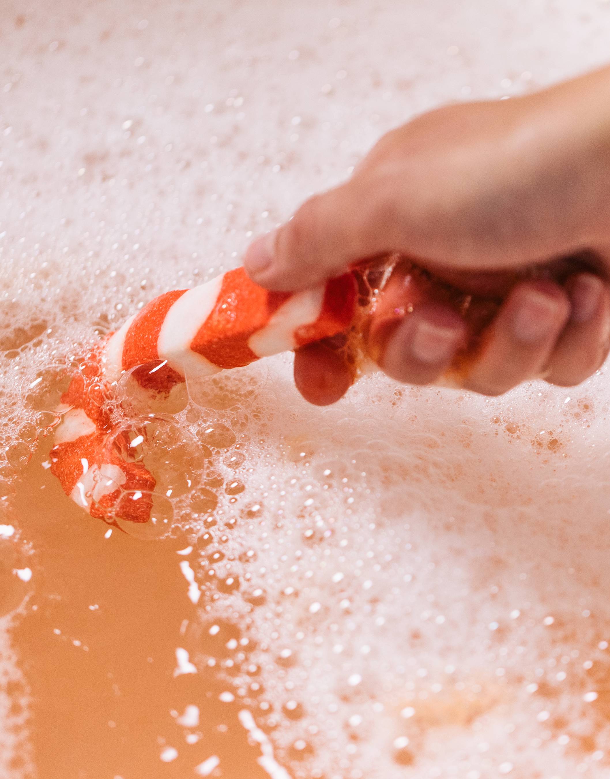 Model holds the Candy Cane bubble bar in the delicate, orange-coloured water covered in a blanket of bubbles. 
