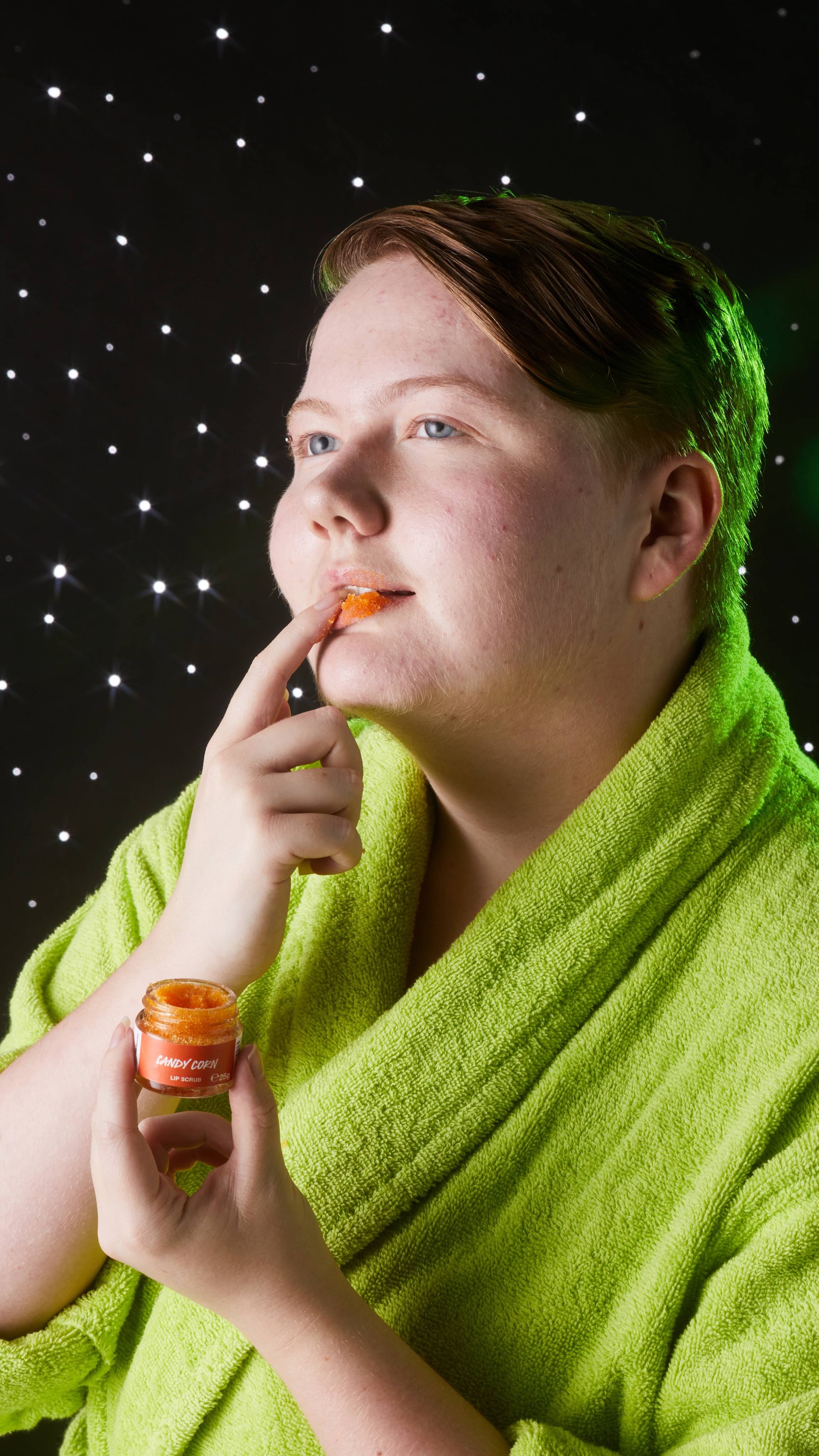 Model is in a neon-green robe on a twinkling background as they gently spread the exfoliating scrub over their lips.