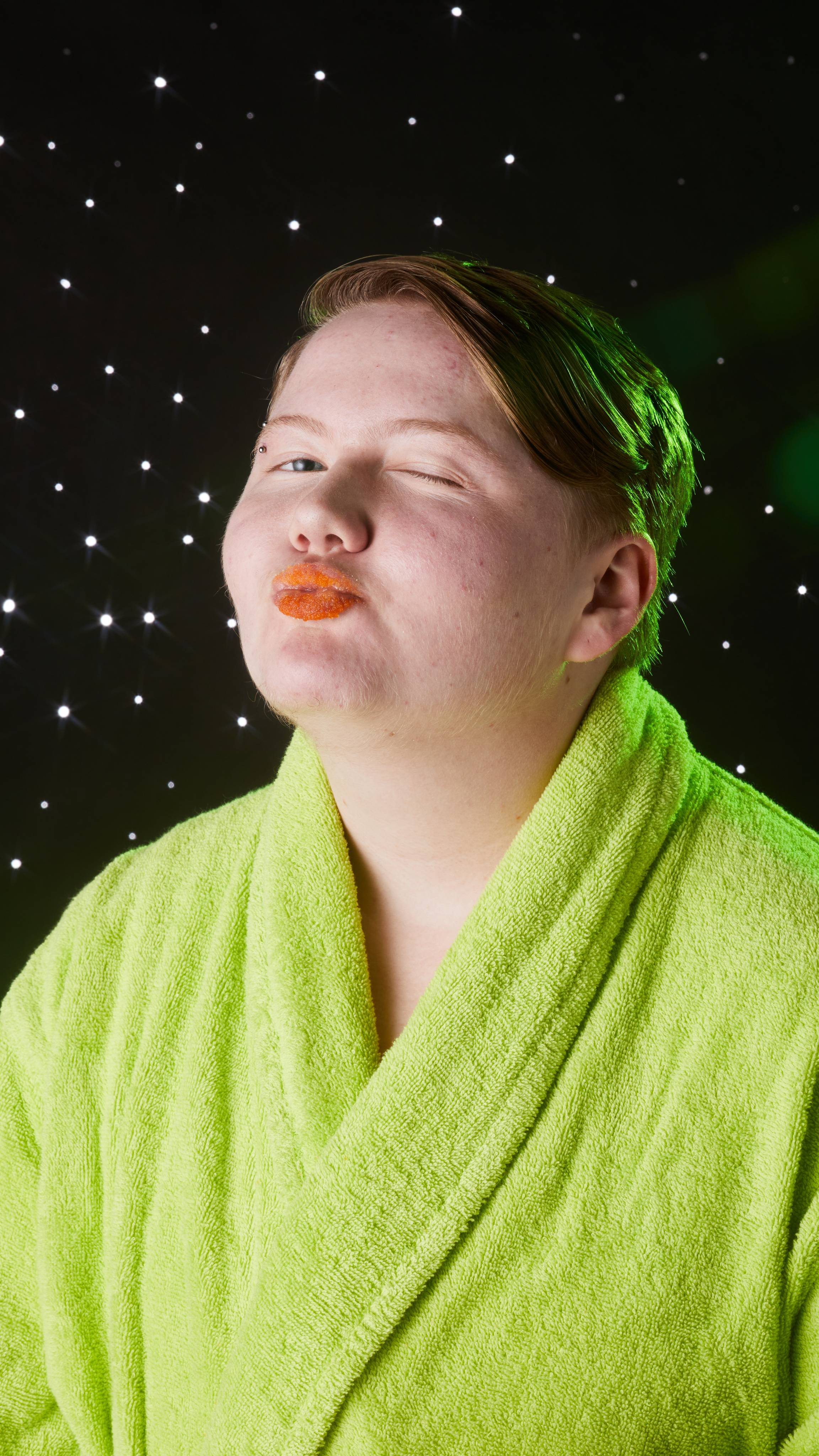 Model is in a neon-green robe as they give a cheeky wink and blow a kiss through orange, sugar-coated lips.