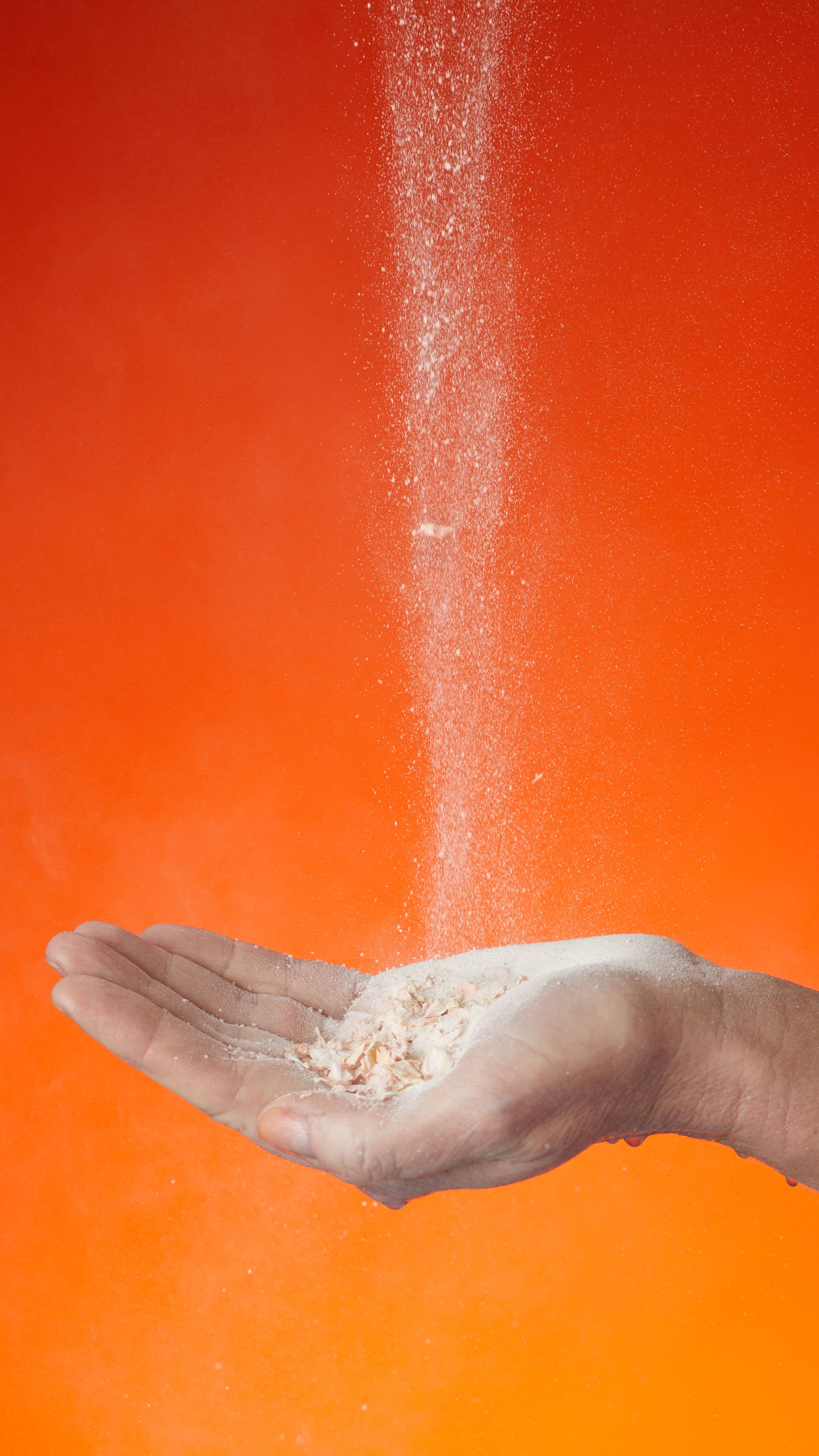 A hand holds the Showder as it is sprinkled. On a dark to light orange gradient background. 