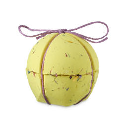 Careys Secret Garden. A large, spherical, yellow bath bomb. The product has visible, dried flowers throughout and is in two halves with a seam around the centre. A pale pink string is crossed and tied in a neat bow to hold the two halves together.
