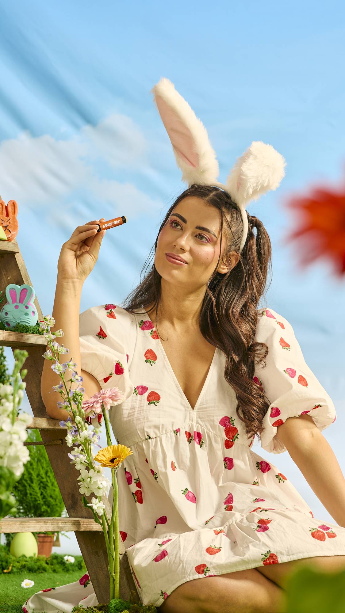 The model is sitting in a white dress with strawberry decoration and they are wearing a bunny ear headband as they hold the Carrot Stick lip balm. 