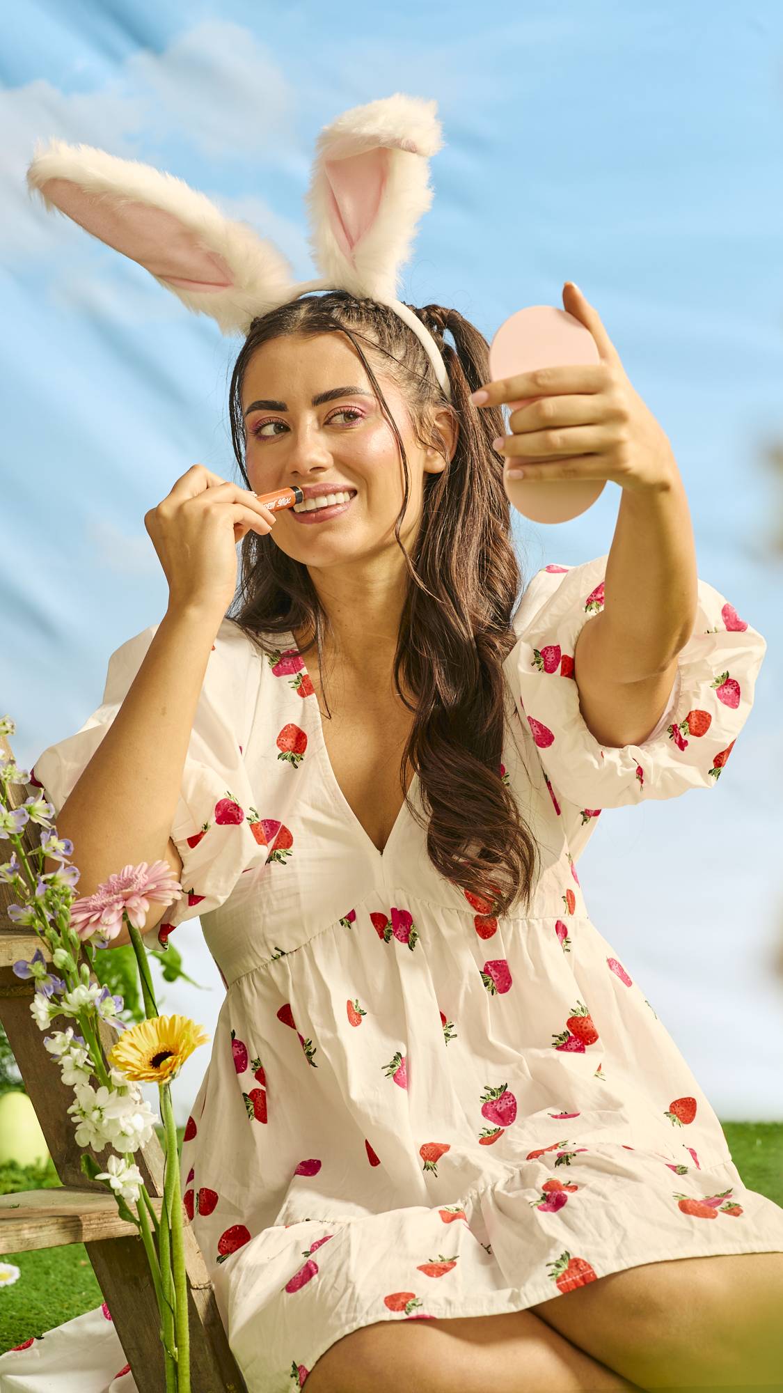 The model is sitting in a white dress with strawberry decoration and they are wearing a bunny ear headband as they apply the Carrot Stick holding a handheld mirror. 