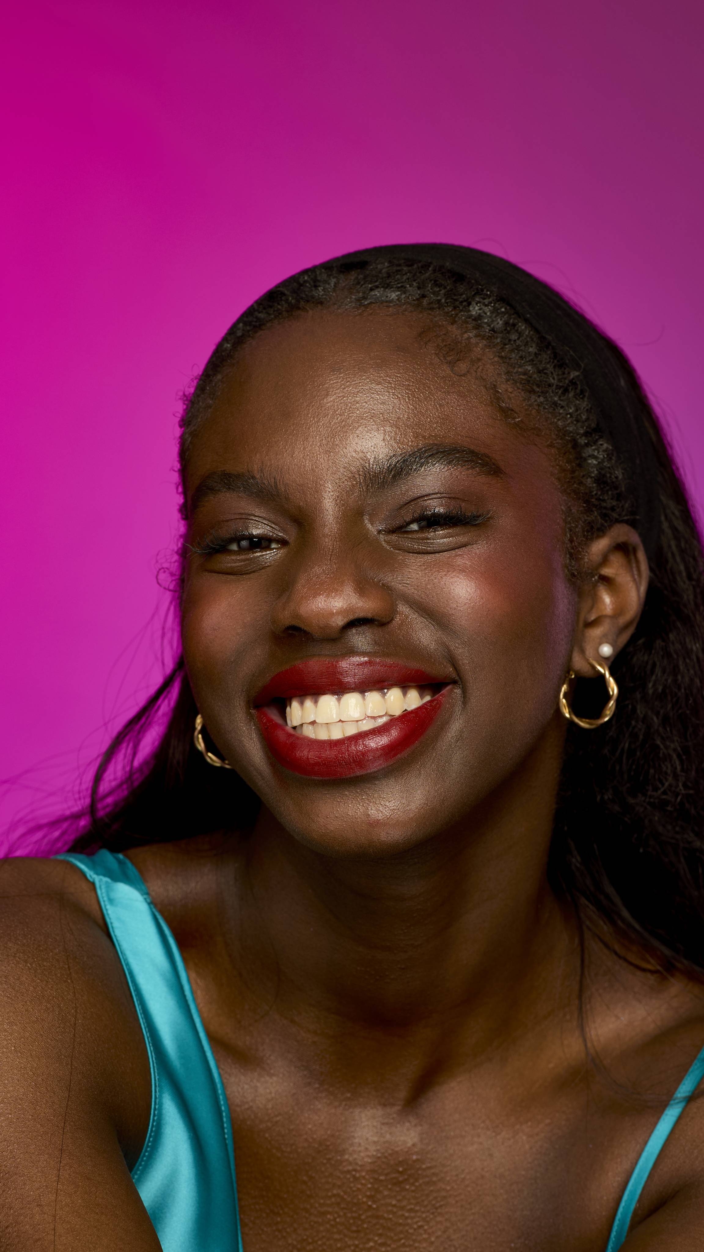 A smiling person with deep skin tone and long, straight dark hair is shown from the chest up, wearing a bright teal top, on a vibrant magenta background. They are wearing the velvety, burgundy-red Catherine Wheel lipstick.