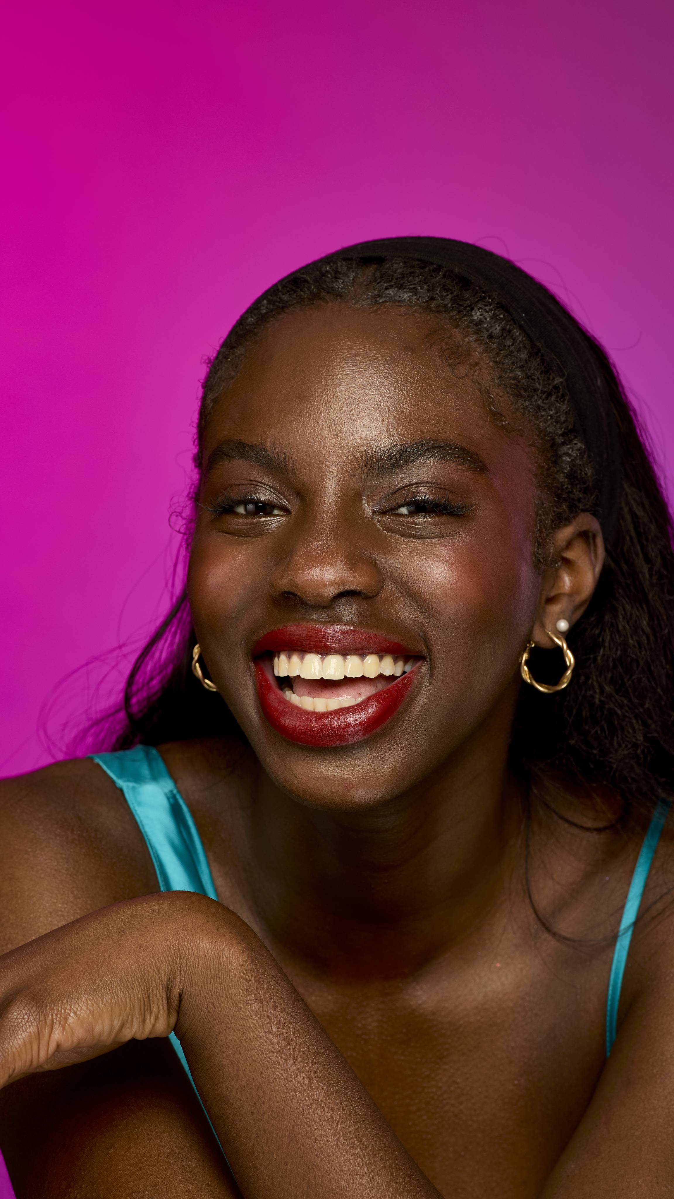A smiling person with deep skin tone and long, straight dark hair is shown from the chest up, wearing a bright teal top, on a vibrant magenta background. They are wearing the velvety, burgundy-red Catherine Wheel lipstick.