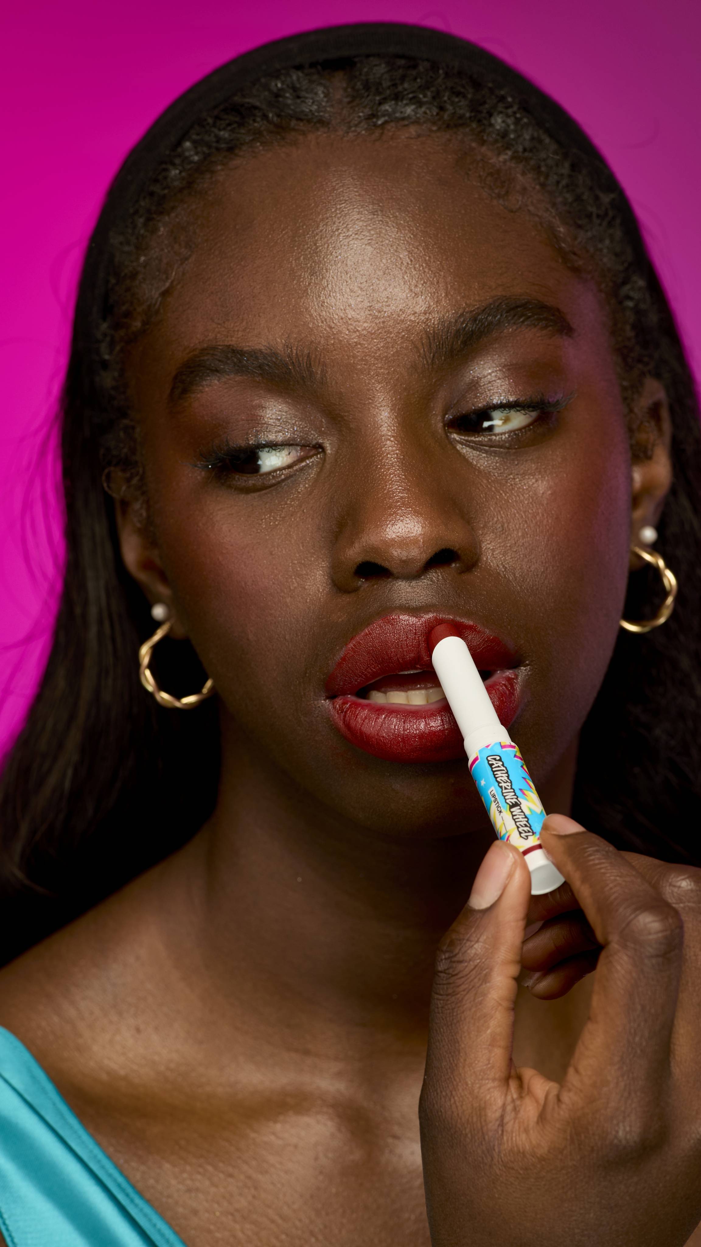 The image shows a model with deep skin tone and long, straight dark hair is shown from the chest up, wearing a bright teal top, on a vibrant magenta background. They are applying the velvety, burgundy-red twist-up Catherine Wheel lipstick.