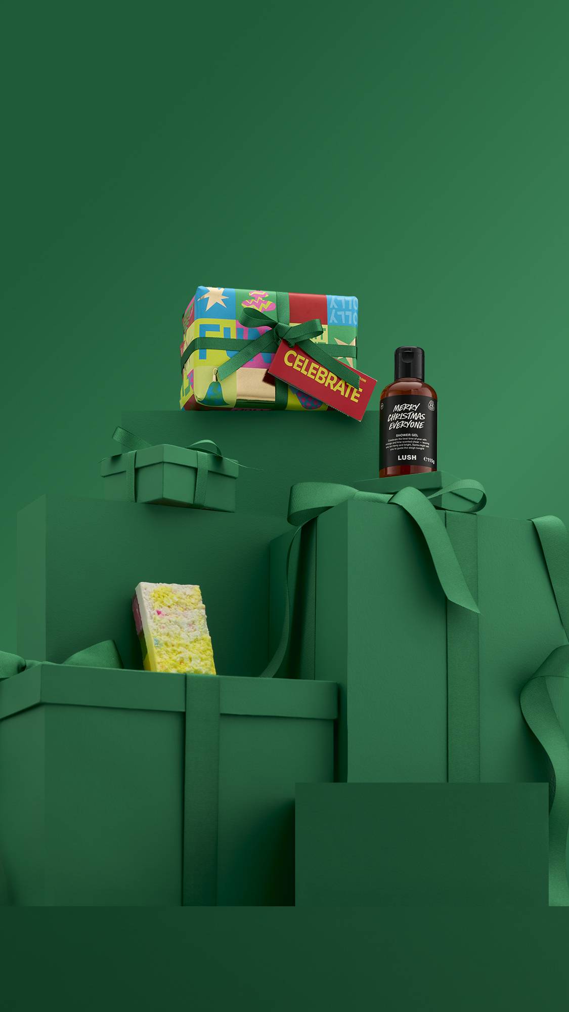 A Lush "Celebrate" gift set. A brightly colored, pop-art-style gift box sits on a dark green background with other wrapped boxes. The set also includes a bottle of "Merry Christmas Everyone" shower gel and a white-and-yellow solid bath product.